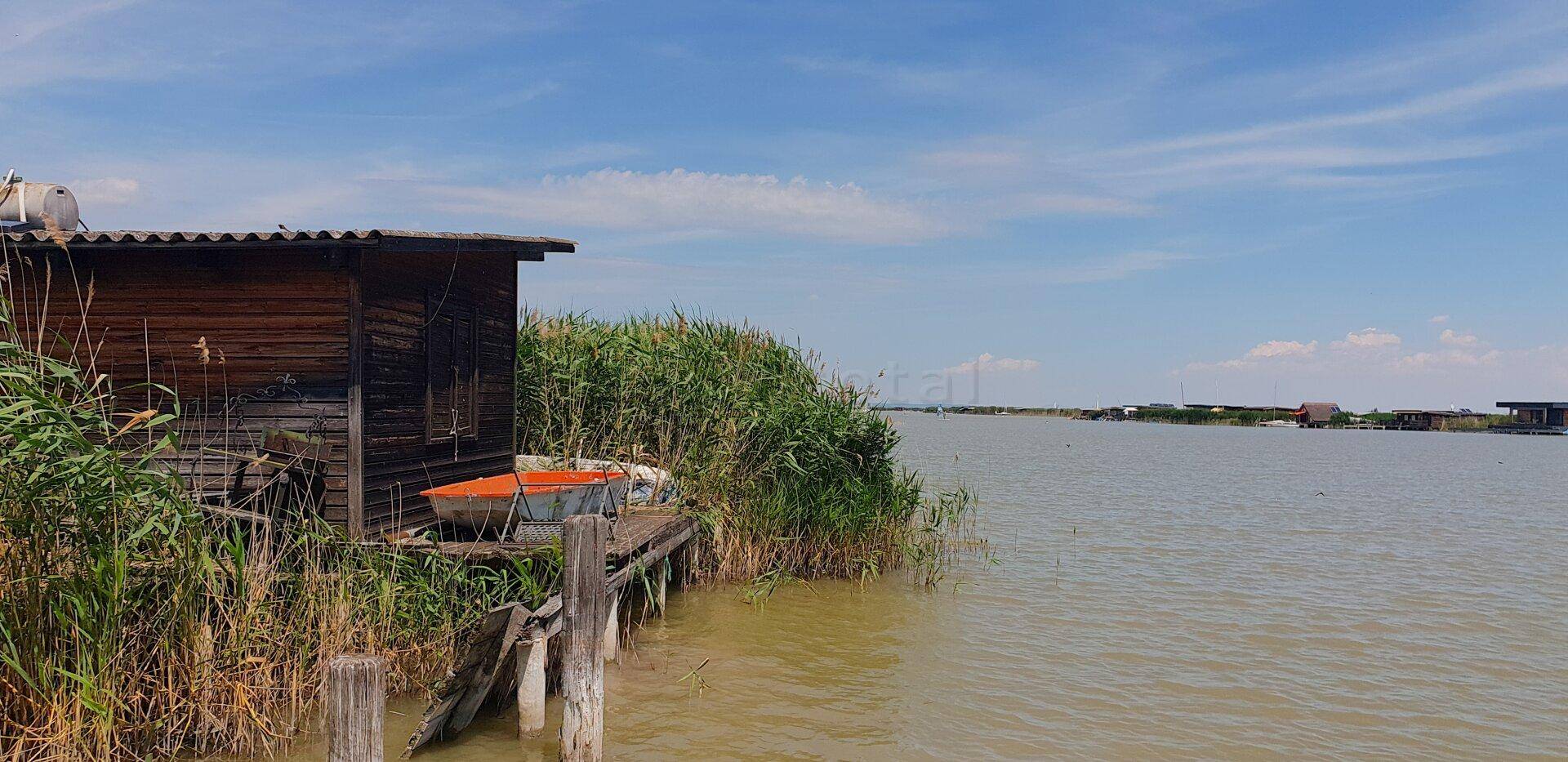 Rustikale Holzhütte direkt am Wasser, eingebettet in üppiges Schilf unter blauem Himmel.