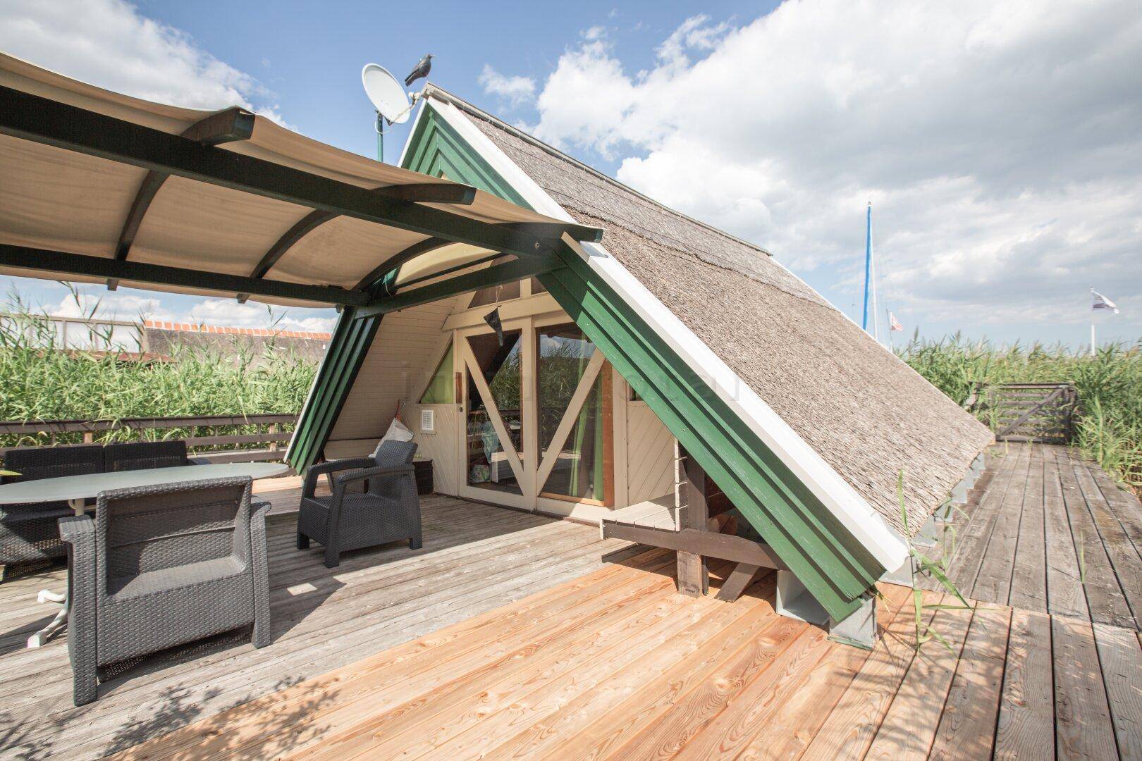 Holzterrasse mit Sitzgelegenheiten und Überdachung vor einem Haus mit Reetdach und Schilfblick.