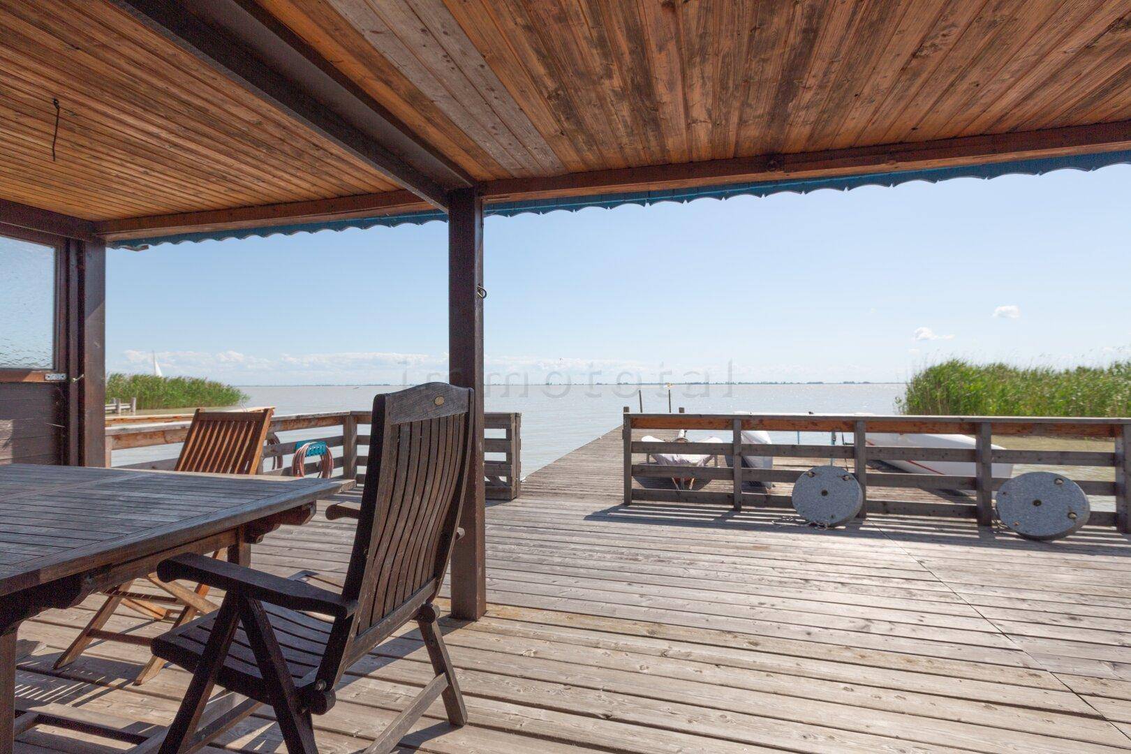 Überdachte Holzterrasse mit Seeblick und Gartenmöbeln unter blauem Himmel.