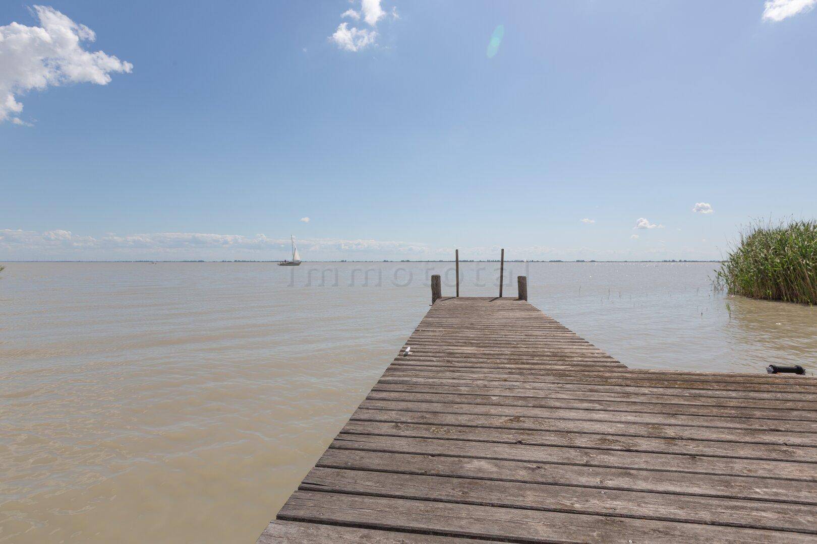 Langer Holzsteg ragt in den See, mit einem Segelboot am Horizont.