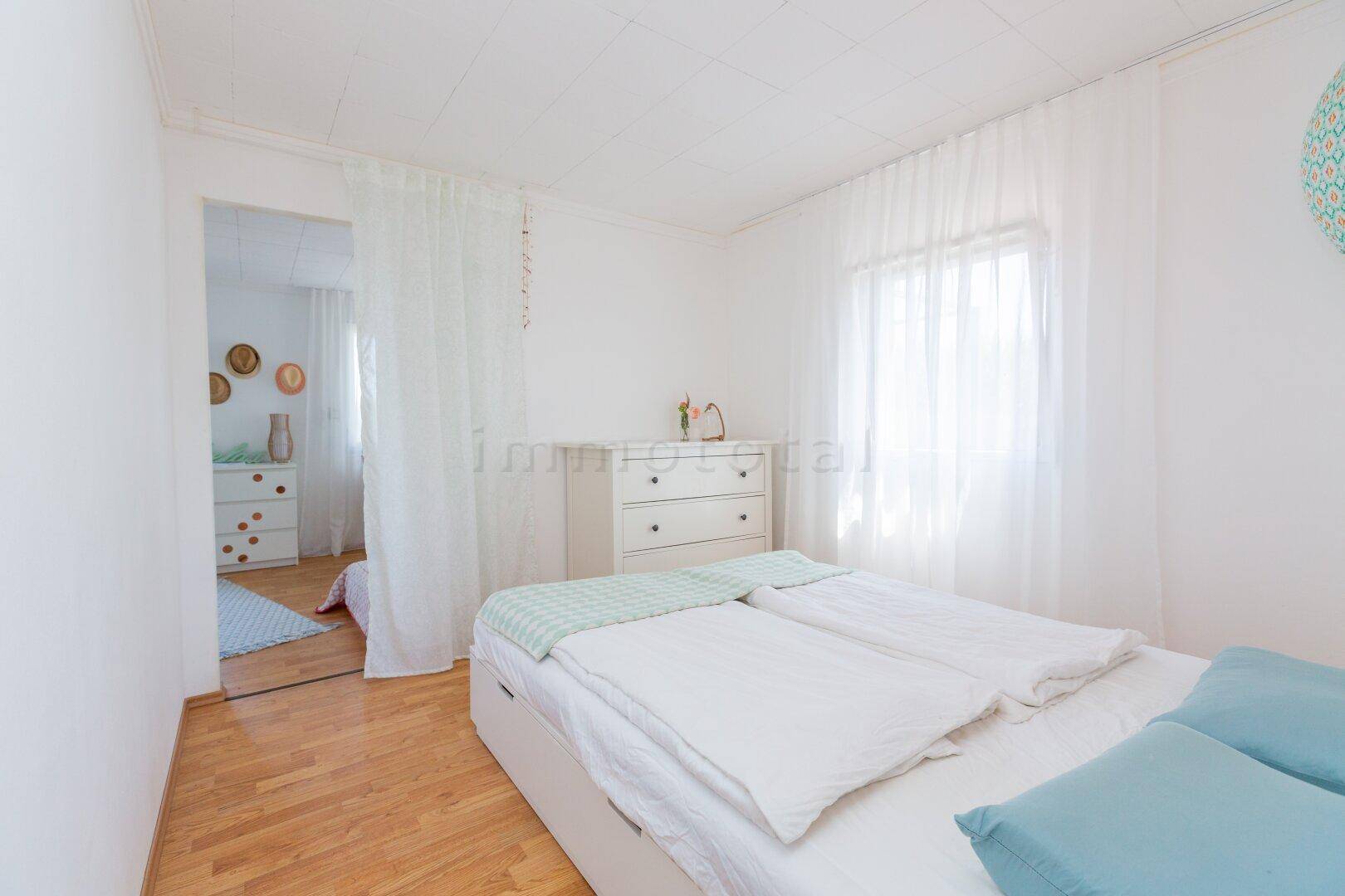 Gemütliches Schlafzimmer mit Doppelbett, weißen Vorhängen und hellem Holzboden.