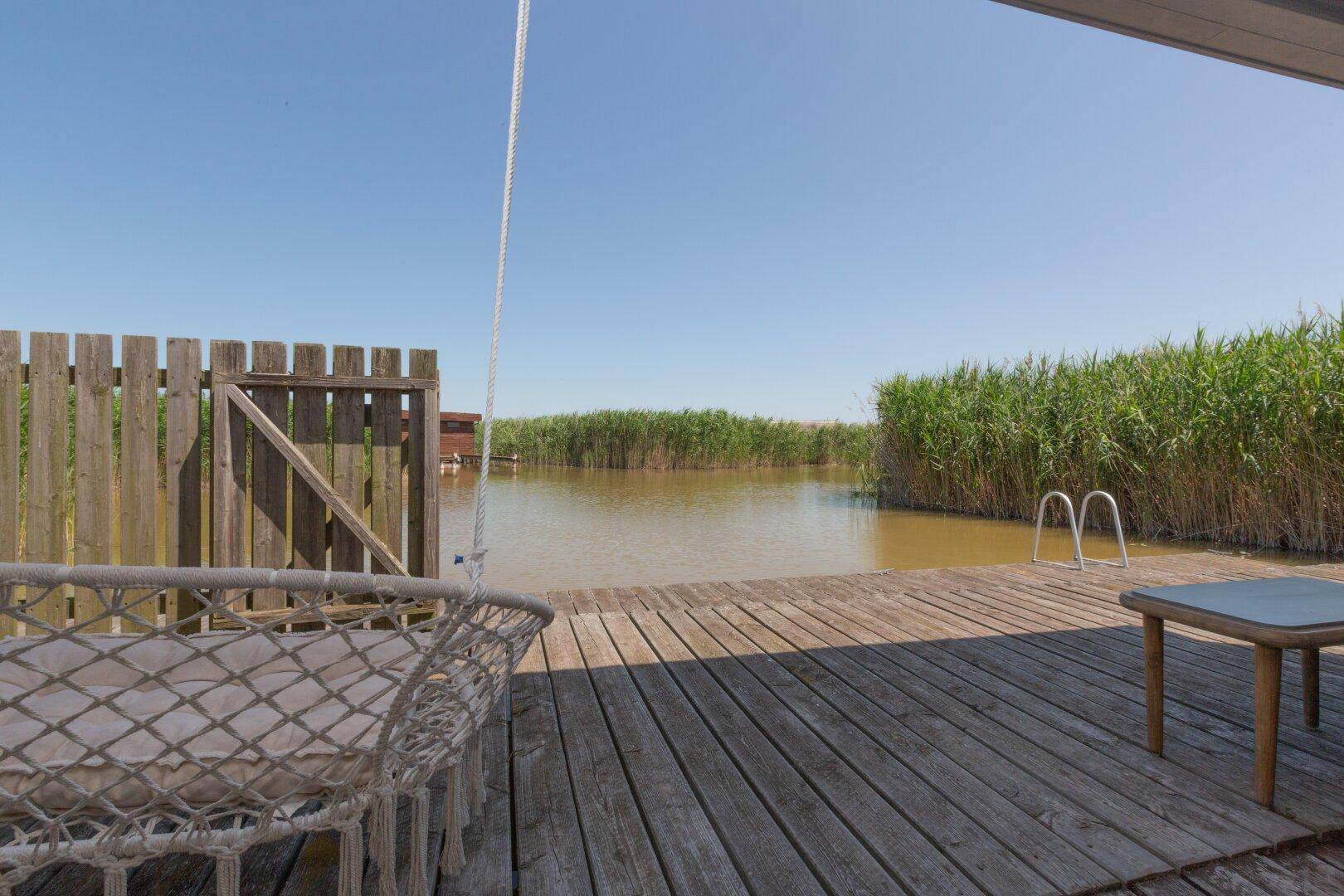 Holzterrasse mit Hängeschaukel und direktem Zugang zum Wasser, umgeben von Schilf.