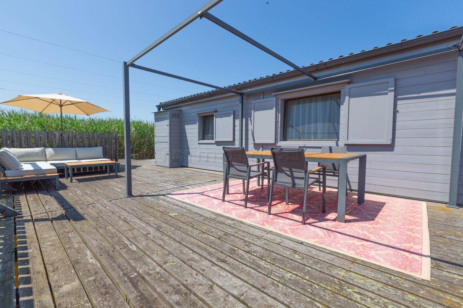 Geräumige Holzterrasse mit Esstisch, Lounge-Möbeln und Sonnenschirm, umgeben von Natur.