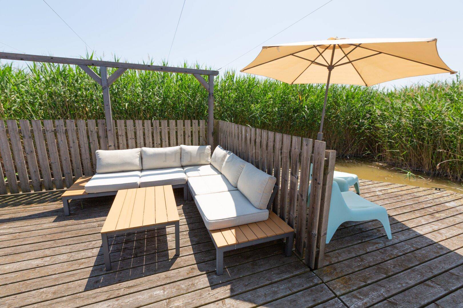 Gemütlicher Lounge-Bereich auf der Holzterrasse mit Sofa und Sonnenschirm, umgeben von Schilf.