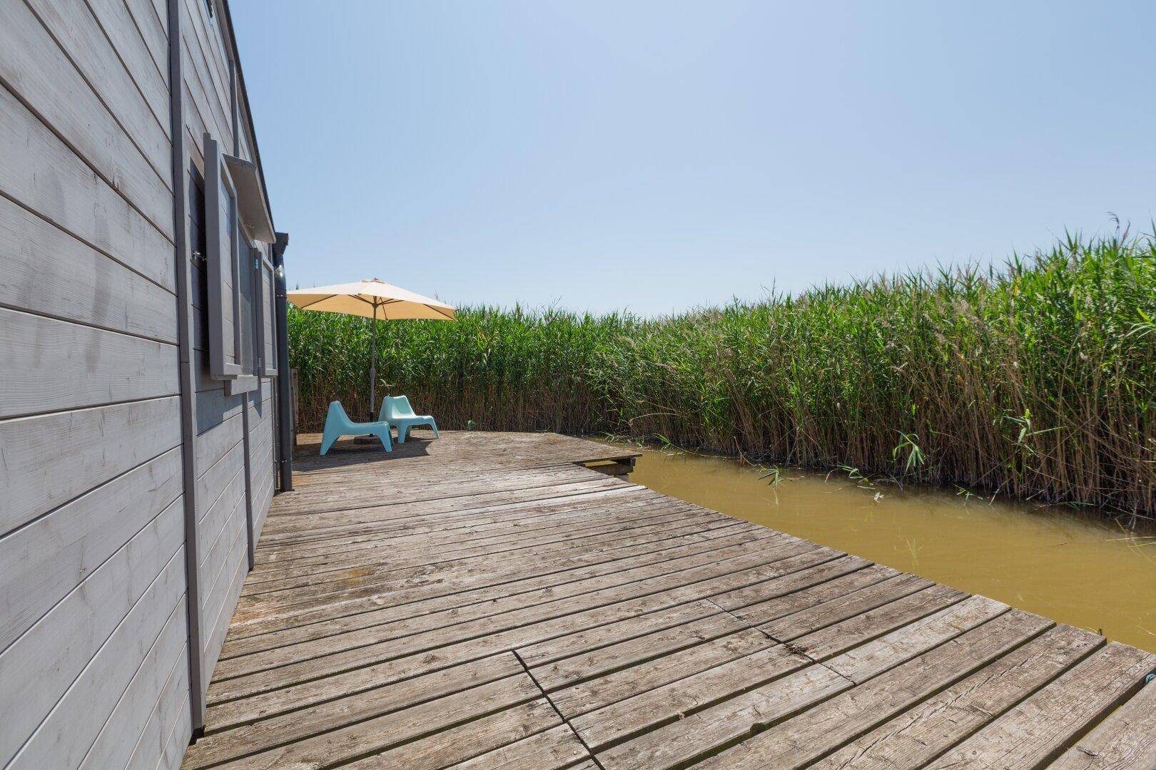 Langer Holzsteg entlang der Hausseite, umgeben von Schilf und Wasser.