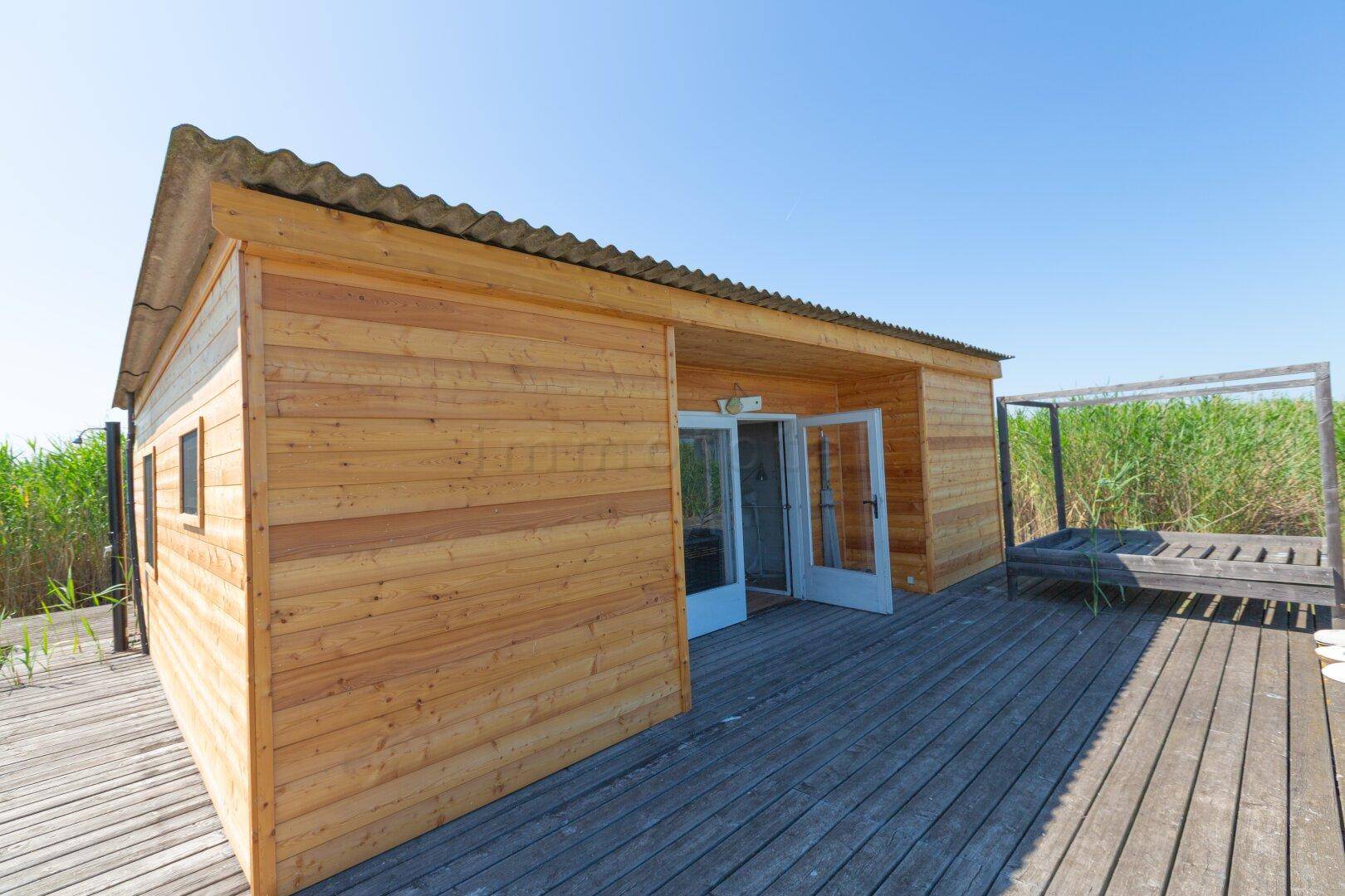 Holzhaus mit großer Holzterrasse und Zugang zum Wasser, umgeben von Schilf.
