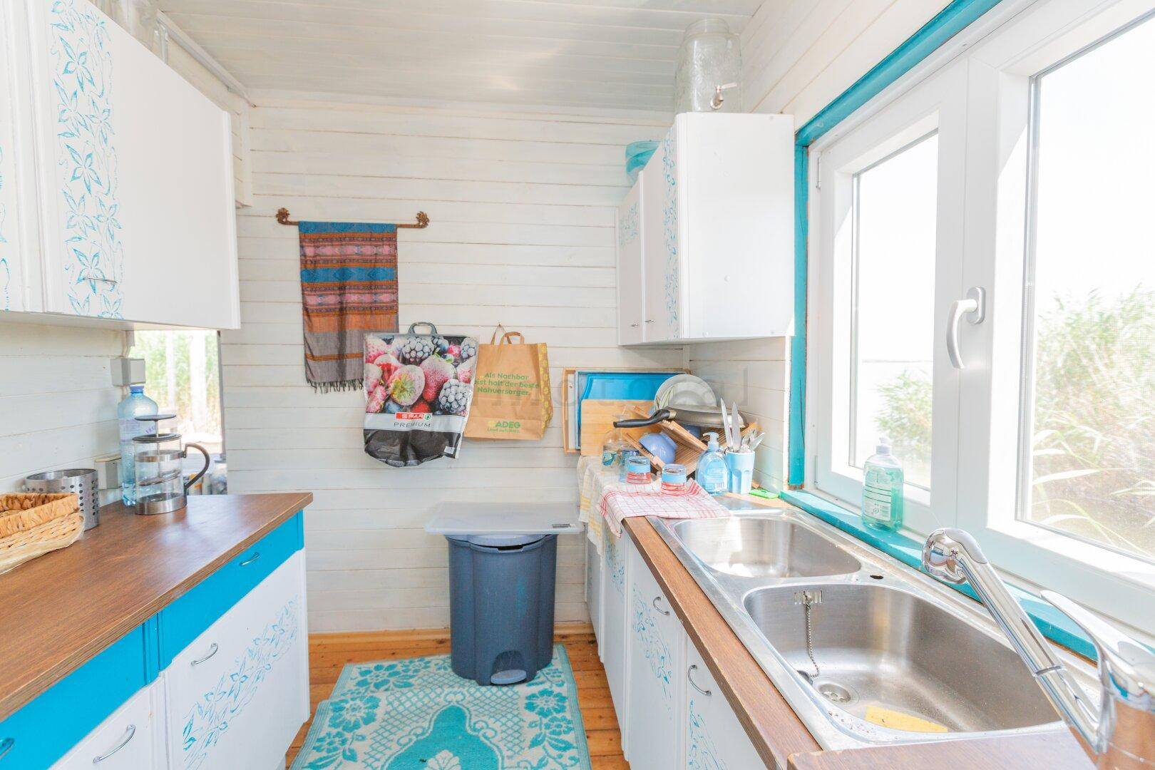 Helle Küche mit Fenster, Doppelspüle und blauen Akzenten, umgeben von weiß getäfelten Wänden.