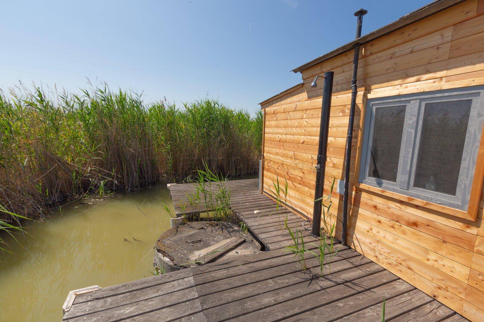 Seitliche Holzterrasse mit Außendusche und Blick auf das Wasser und die Schilflandschaft.