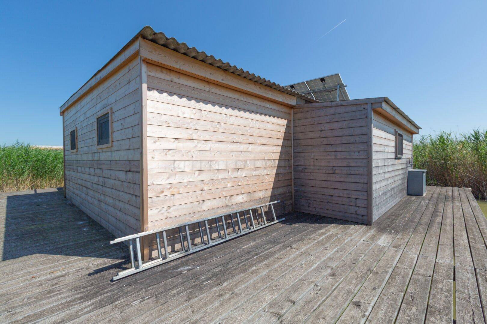 Rückseite des Holzhauses mit Solarpaneelen und einer Leiter auf der Holzterrasse.