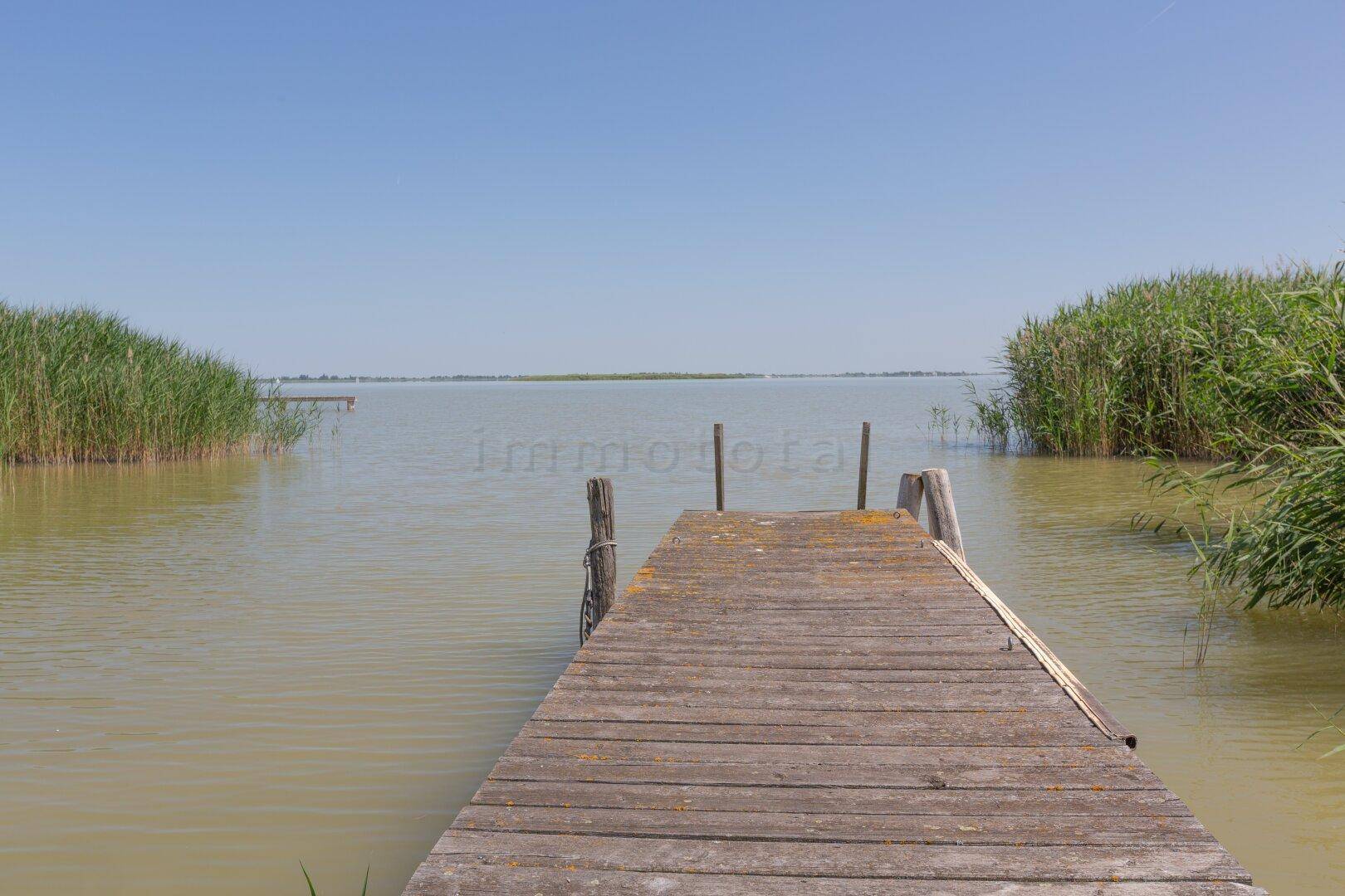 Langer Holzsteg führt in die weite Wasserlandschaft, umgeben von Schilf unter blauem Himmel.