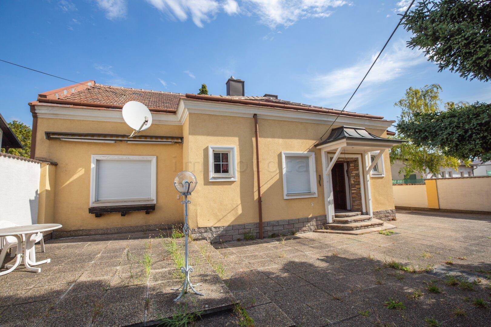 Gelbes Einfamilienhaus mit gepflastertem Vorplatz und einer kleinen Terrasse mit Gartenmöbeln.
