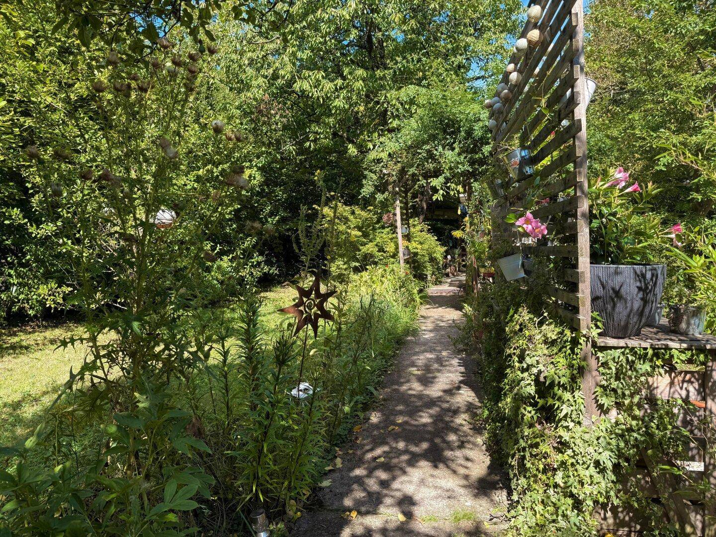 Sonniger Gartenweg mit blühenden Pflanzen, dekorativen Elementen und einer Holzpergola im Hintergrund.