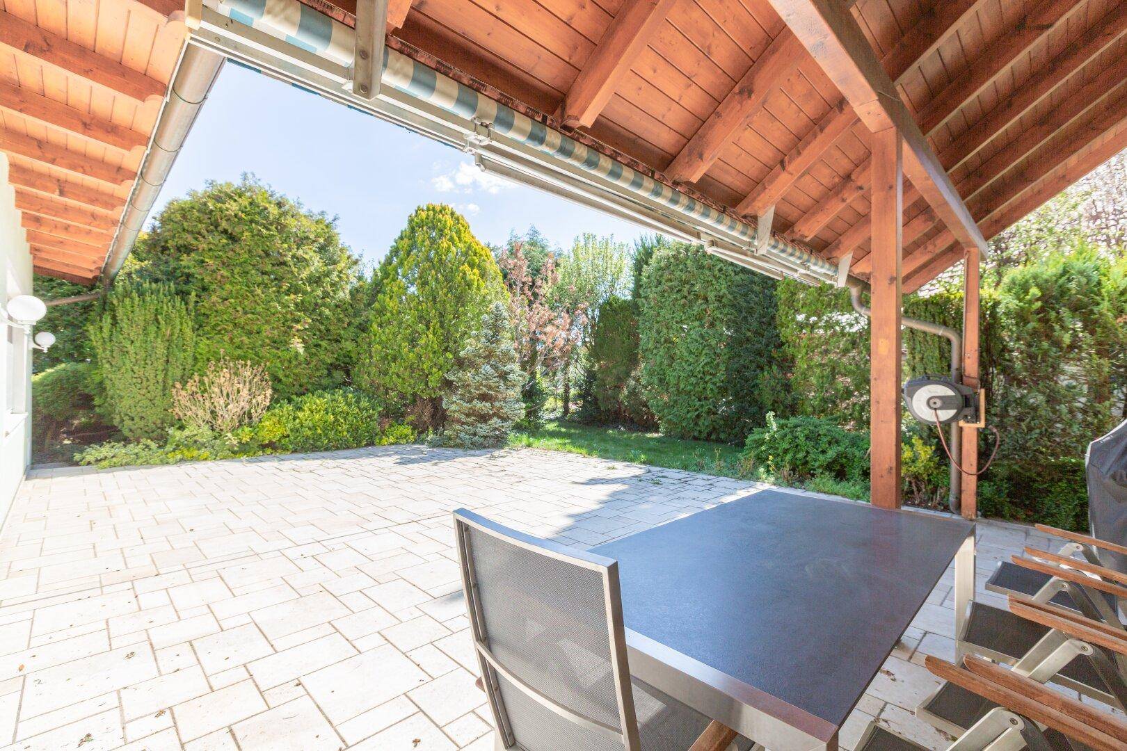 Überdachte Terrasse mit Esstisch und Stühlen, umgeben von üppigem Grün.