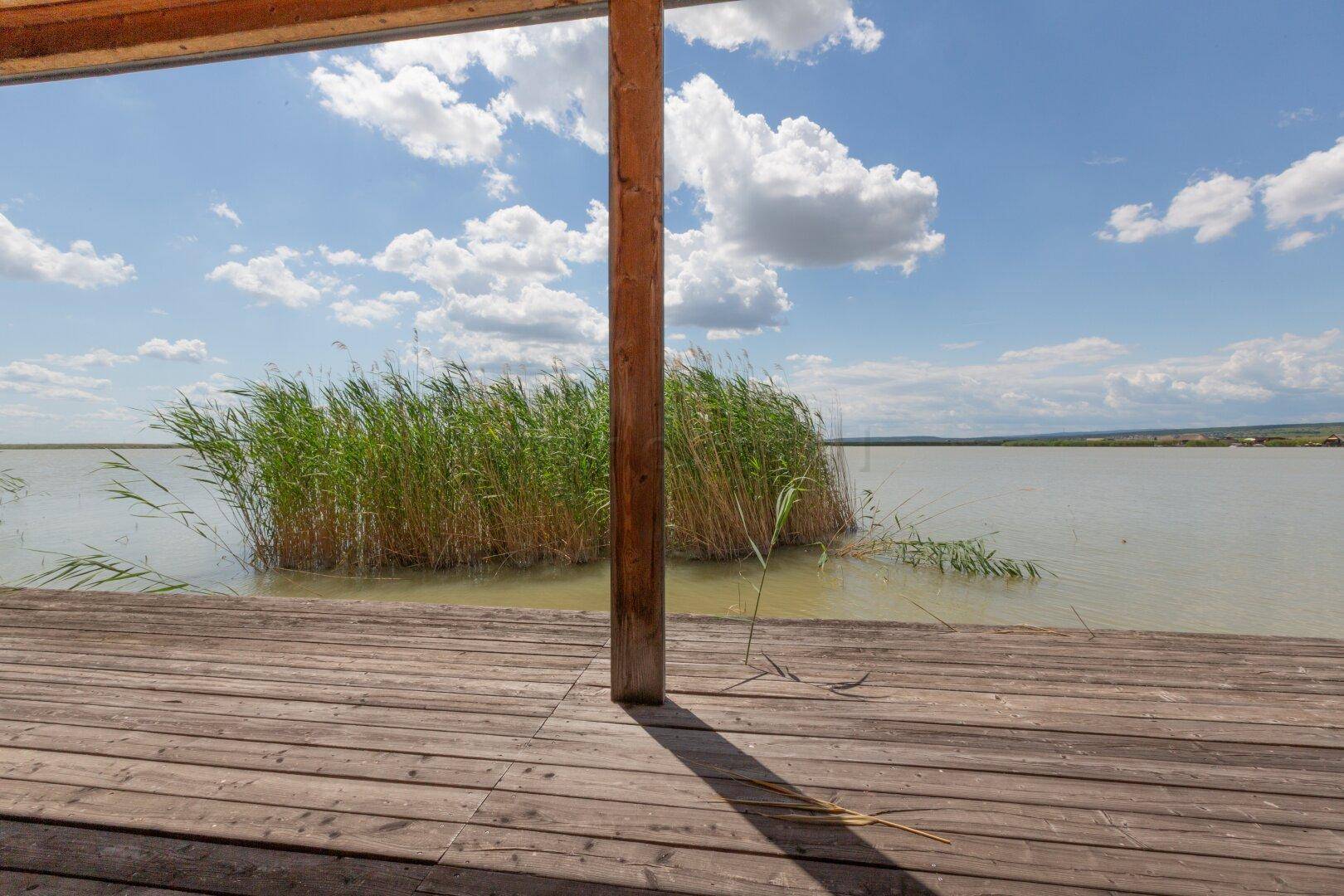 Holzterrasse mit Blick auf das ruhige Wasser des Sees und die umgebende Schilflandschaft.