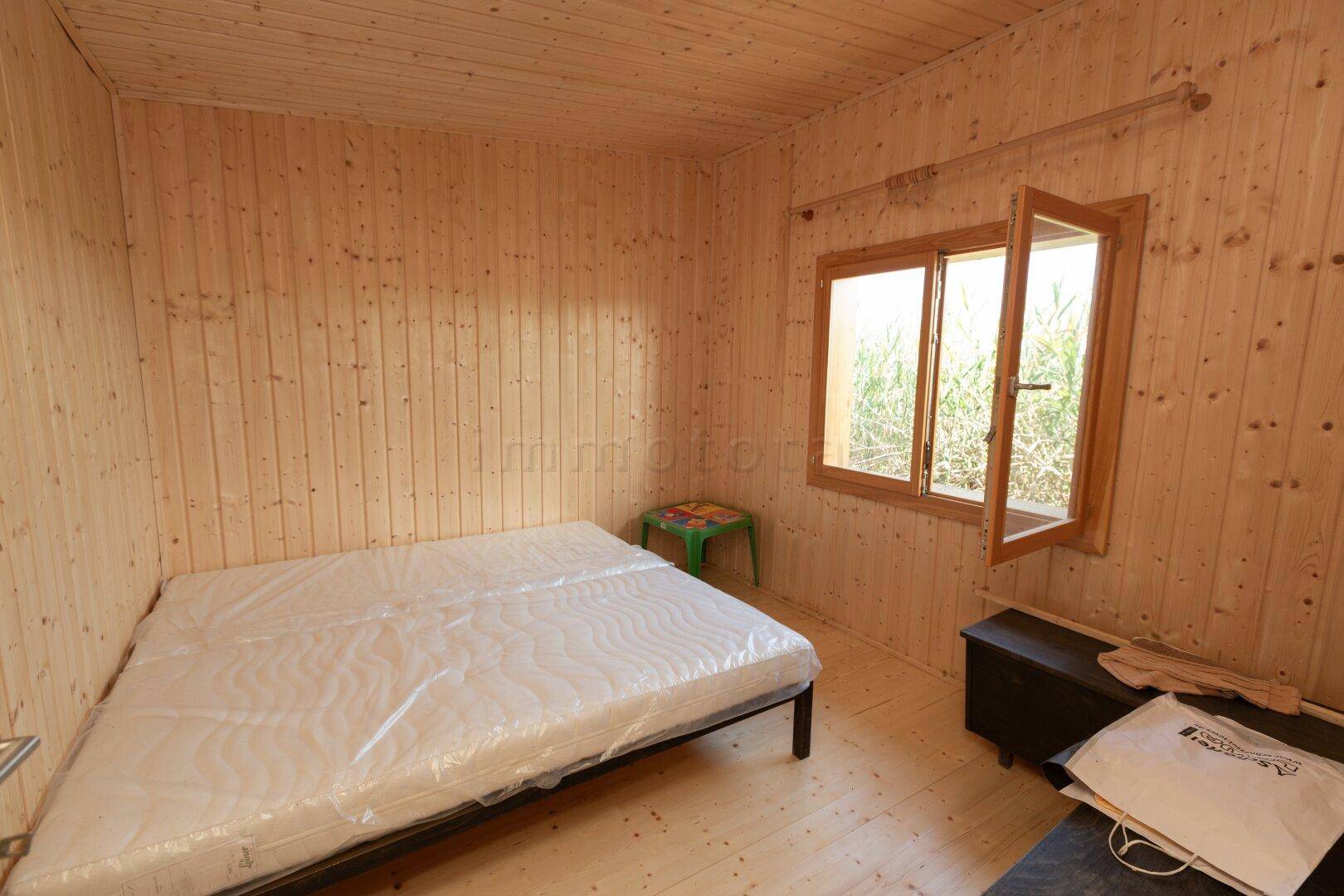 Schlafzimmer mit Holzvertäfelung, einem Bett mit Matratze und einem Fenster mit Blick ins Grüne.