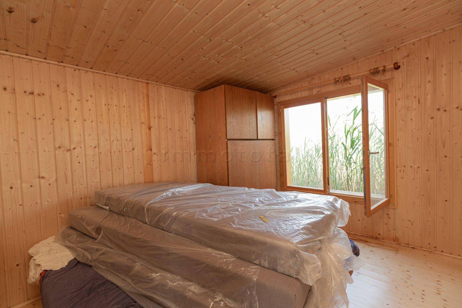 Weiteres Schlafzimmer mit Holzvertäfelung, gestapelten Matratzen und einem Fenster zur Natur.