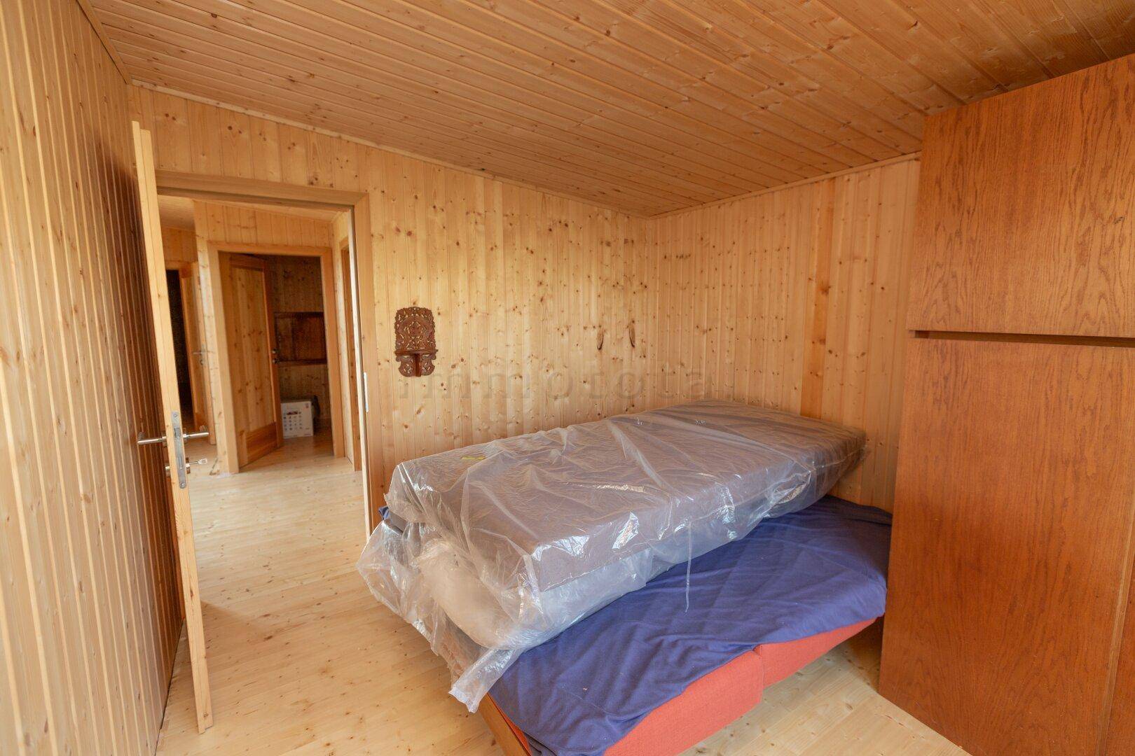 Schlafzimmer mit Holzwänden und -boden, Blick auf den Flur und gestapelte Matratzen.