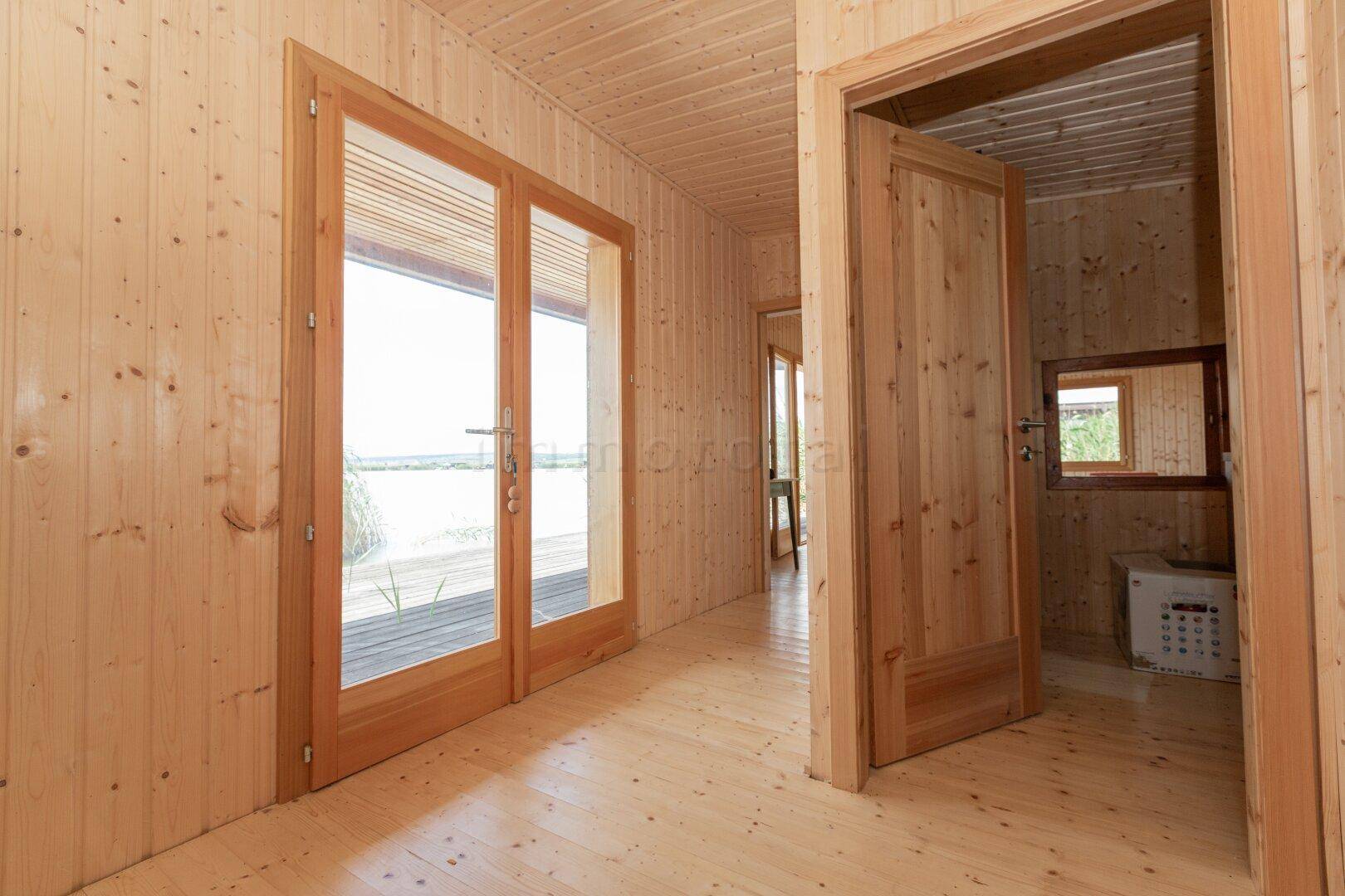 Flur mit Holzboden und -wänden, der zum Wohnbereich führt und Seeblick durch Glastüren bietet.
