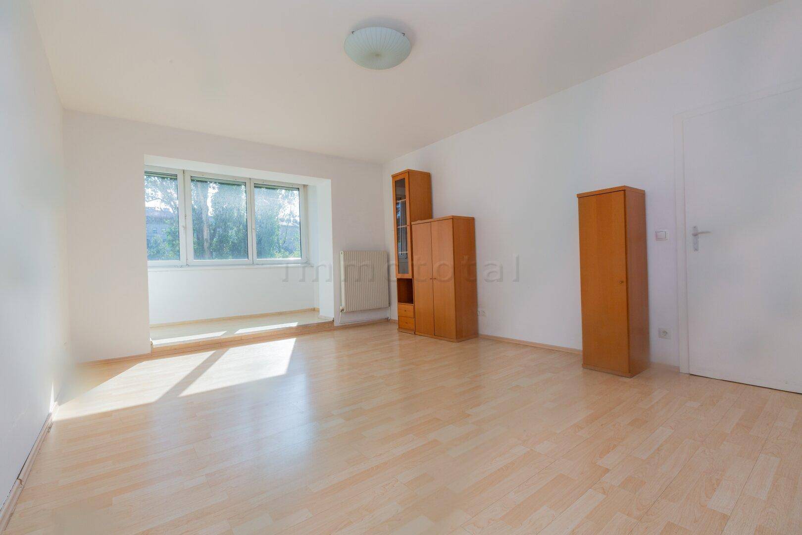 Helles, leeres Zimmer mit Laminatboden und großem Fenster, das viel Tageslicht hereinlässt.