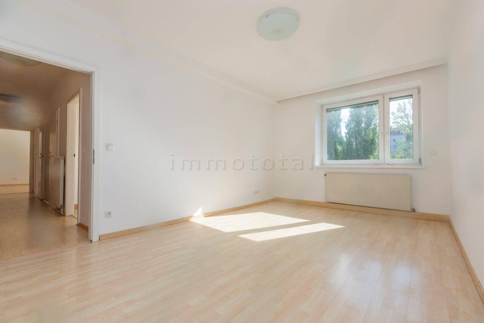 Leeres Schlafzimmer mit hellem Laminatboden und einem Fenster, das Sonnenlicht hereinlässt.