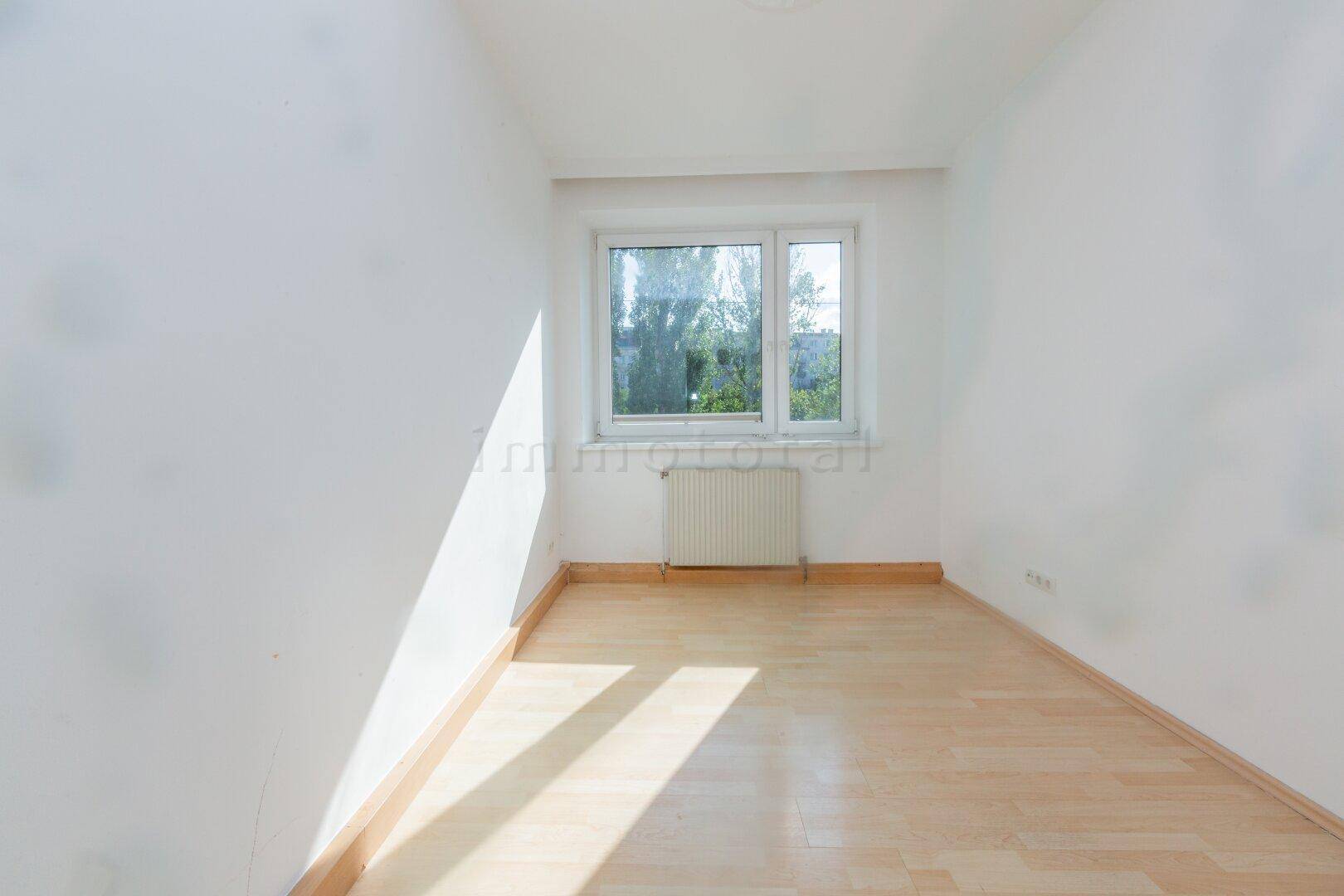 Leeres Zimmer mit Laminatboden und einem Fenster, das den Blick ins Grüne freigibt.