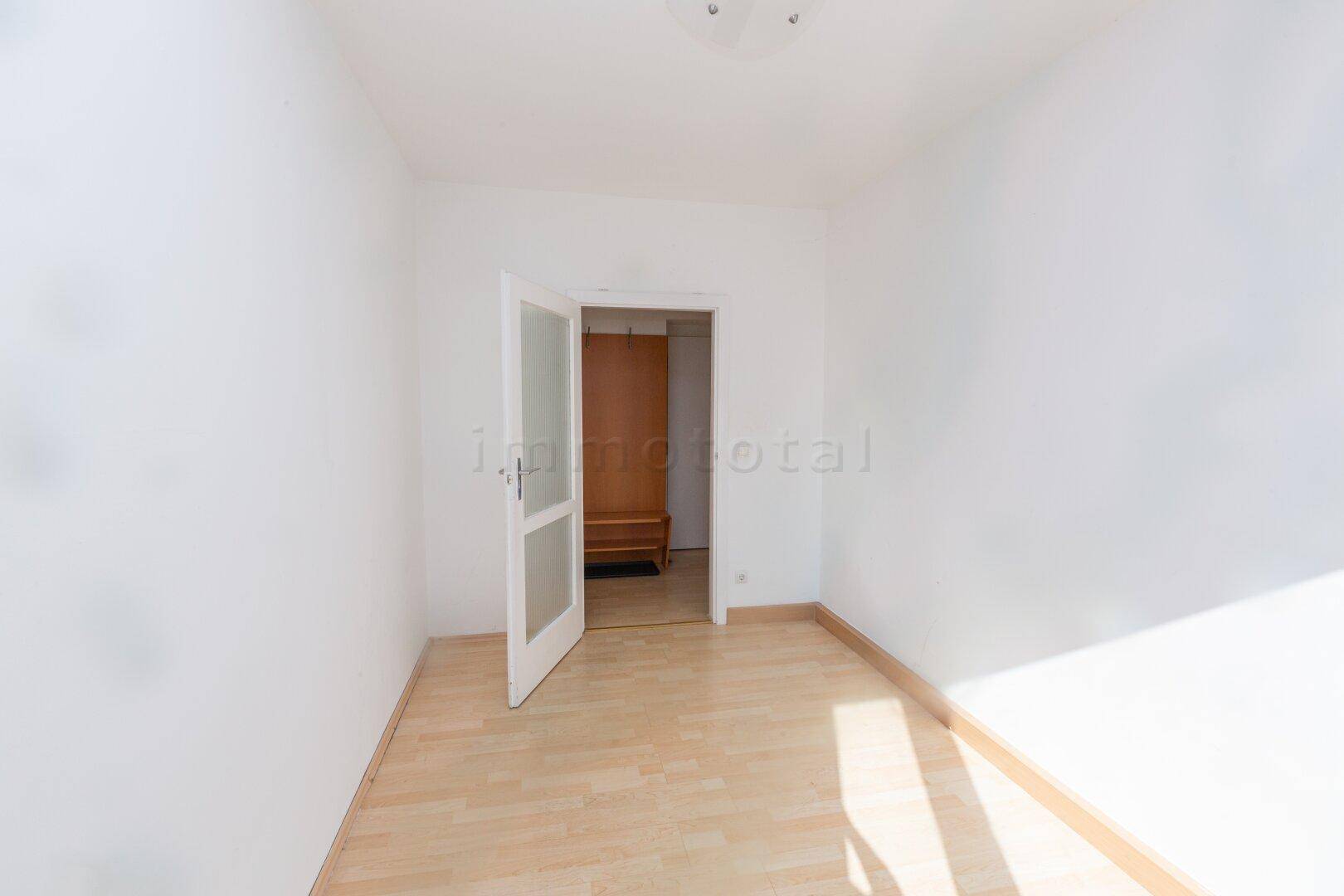 Helles, leeres Zimmer mit Laminatboden und einer Tür, die zu einem weiteren Raum führt.