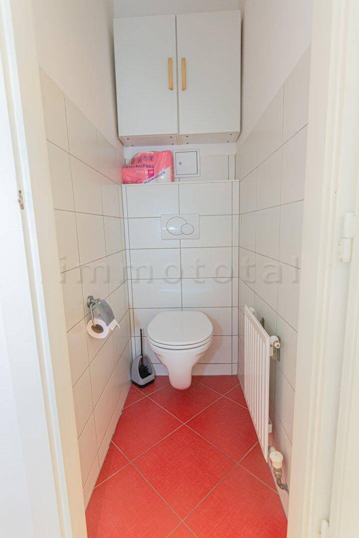 Separate Toilette mit roten Bodenfliesen und weißen Wandfliesen, inklusive Hängeschrank.