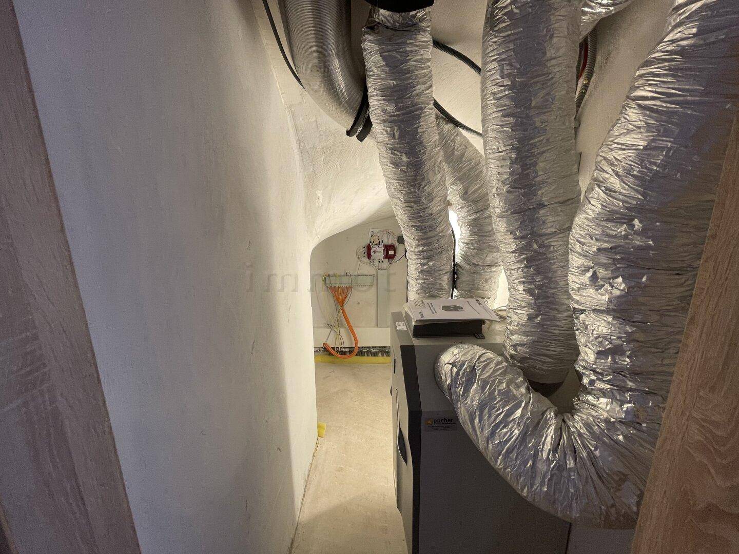 Technischer Raum mit Lüftungsanlage und sichtbaren Leitungen im Kellerbereich.