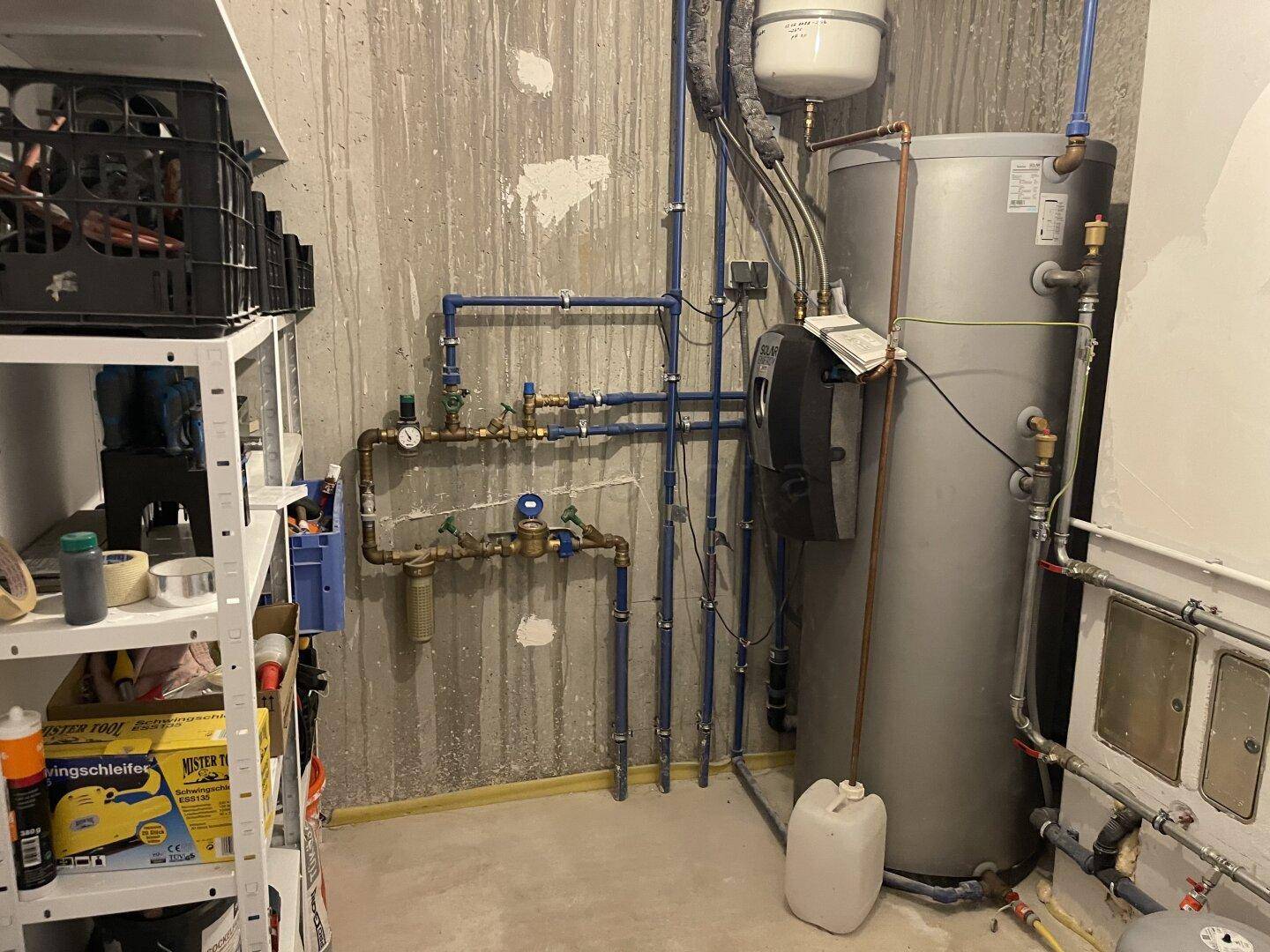 Heizungsraum mit Warmwasserspeicher, Rohrleitungen und Lagerregalen im Untergeschoss.