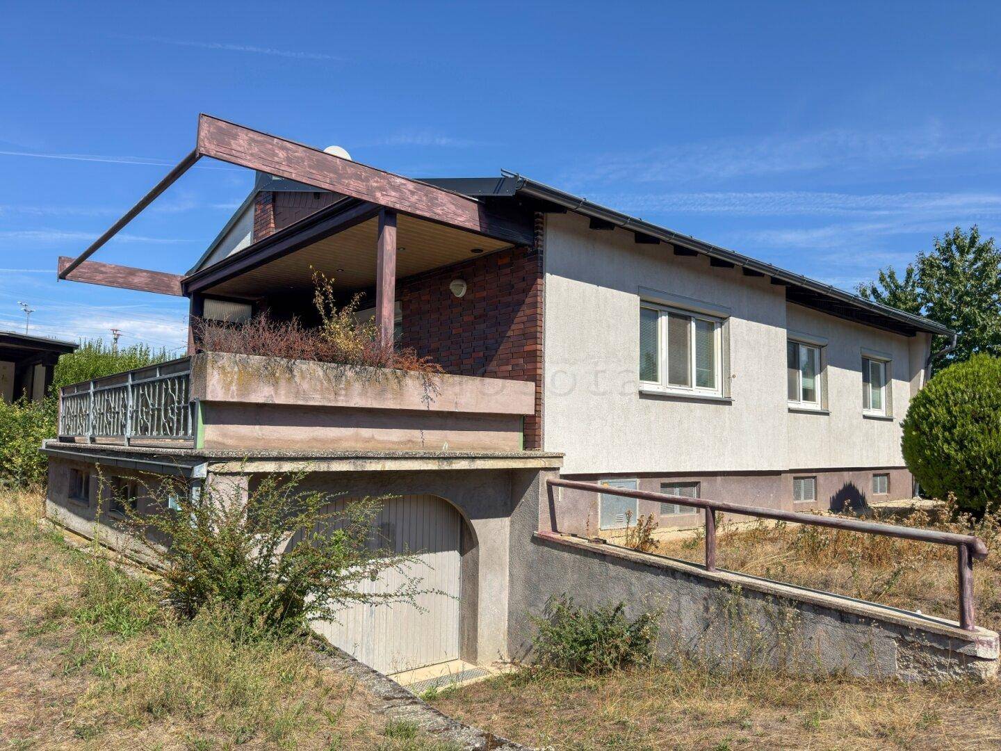 Rückansicht des Hauses mit Garagenzufahrt und Balkon, umgeben von üppiger Vegetation.