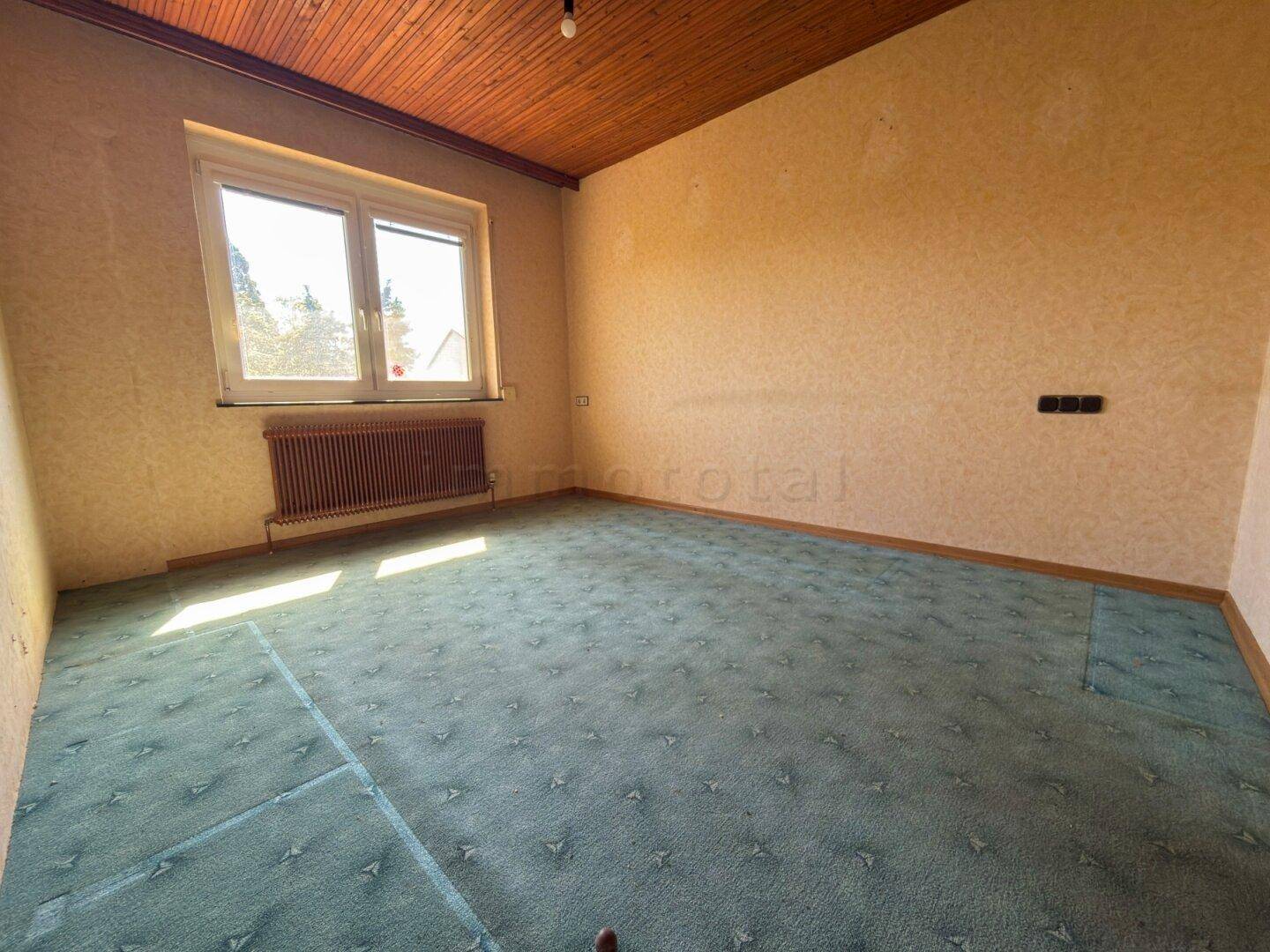 Schlafzimmer mit grünem Teppichboden, Holzdecke und Fenster, renovierungsbedürftig.
