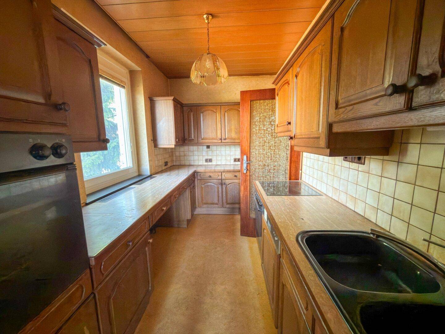 Traditionelle Küche mit Holzschränken, Fliesenrückwand und Fenster, renovierungsbedürftig.