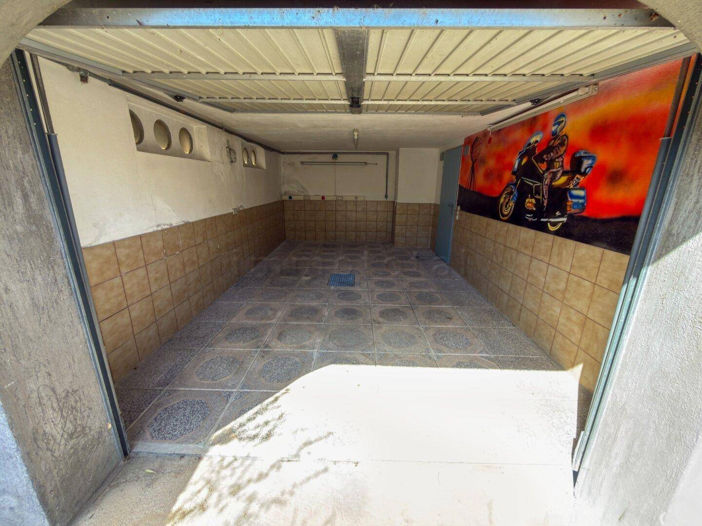 Garage mit Fliesenboden und Wandgemälde, bietet Platz für Fahrzeuge und Hobbys.