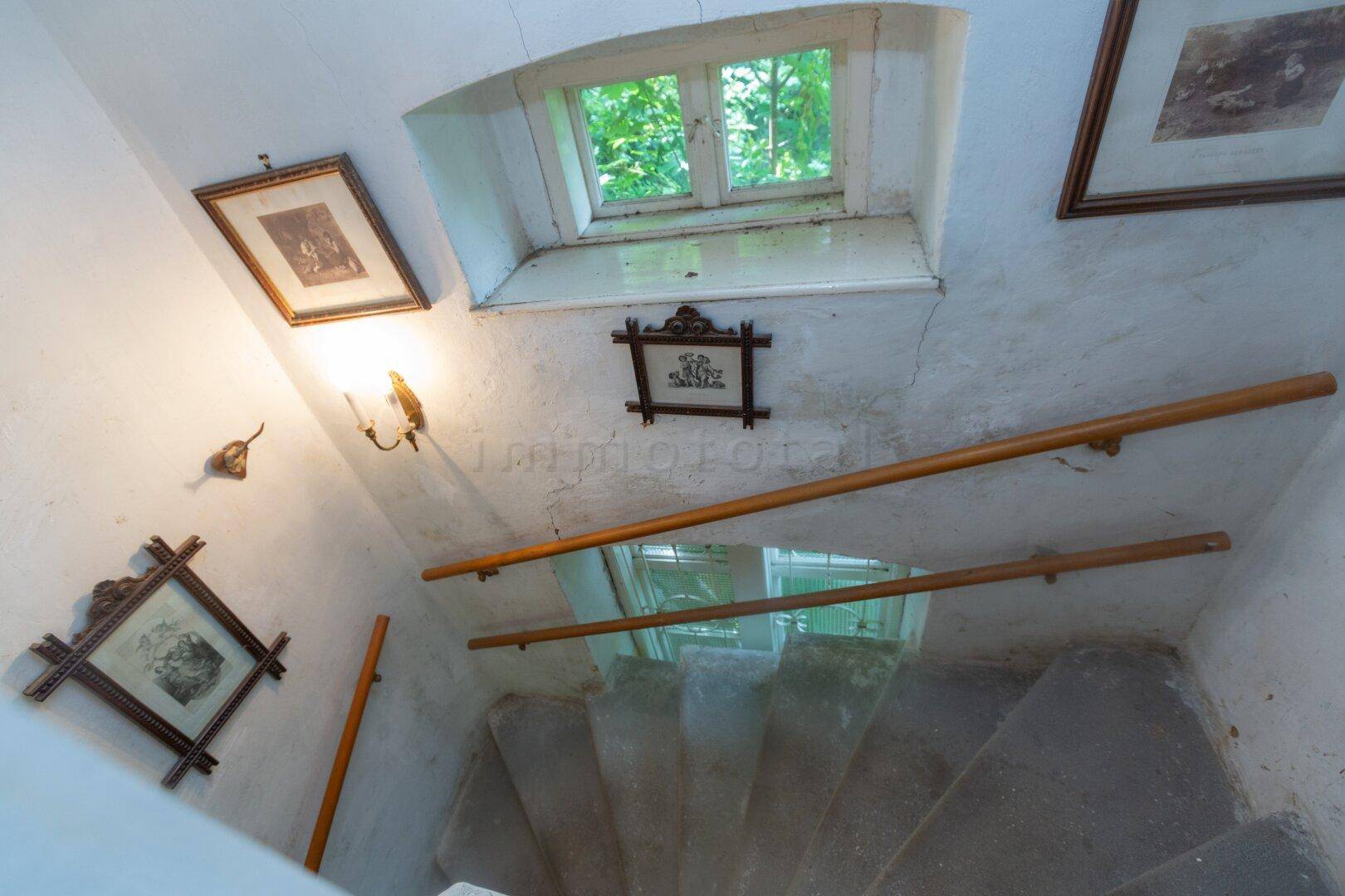 Historisches Treppenhaus mit Holzgeländer, Fenster und gerahmten Bildern an den Wänden.