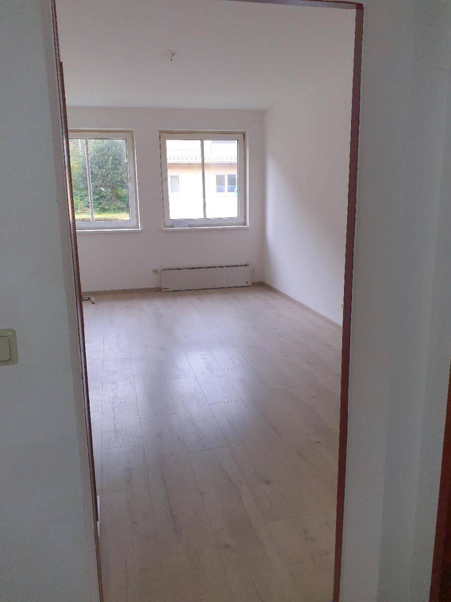 Leeres Schlafzimmer mit hellem Laminatboden und zwei Fenstern, die viel natürliches Licht hereinlassen.