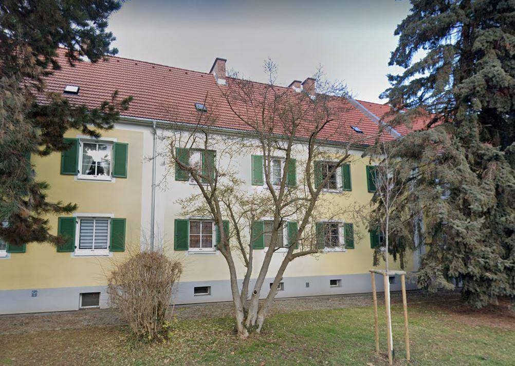 Gelb-weißes Mehrfamilienhaus mit rotem Ziegeldach, grünen Fensterläden und Bäumen im Gartenbereich.