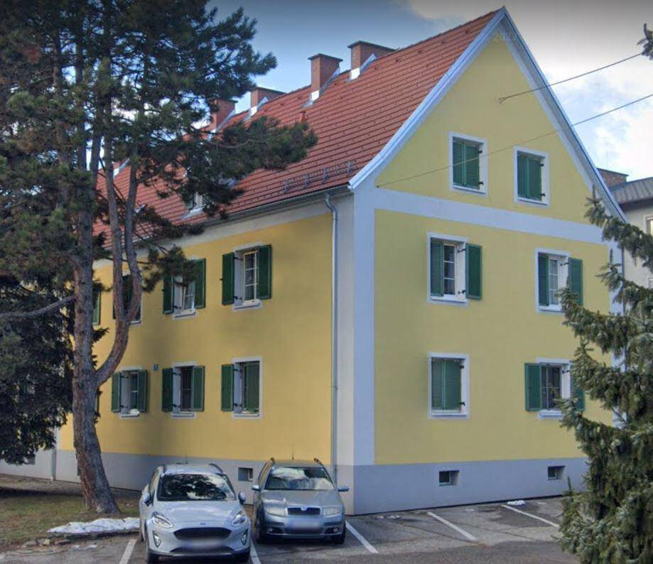 Gelbes Mehrfamilienhaus mit rotem Ziegeldach, grünen Fensterläden und geparkten Autos.