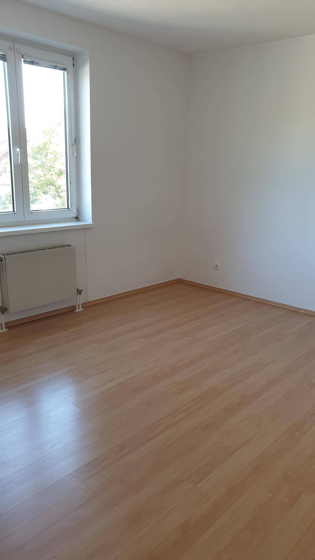 Leeres Zimmer mit Laminatboden und einem Fenster, das Tageslicht spendet.