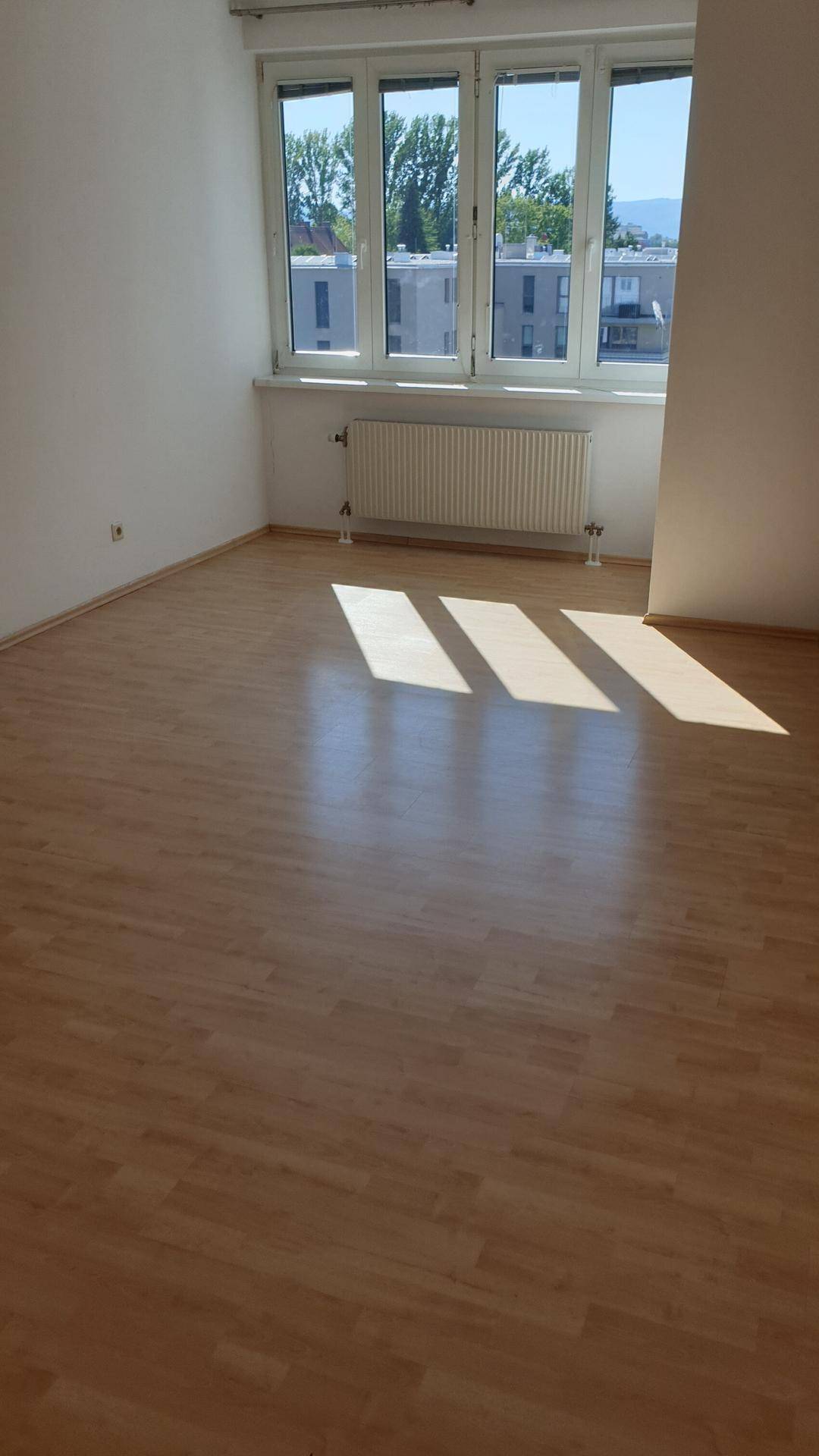 Helles, leeres Zimmer mit Laminatboden und großen Fenstern, die viel Sonnenlicht hereinlassen.