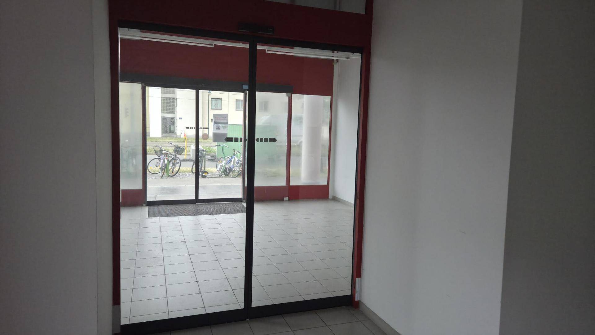 Eingangsbereich mit automatischen Glastüren, Fliesenboden und Blick auf die Straße und Fahrräder.