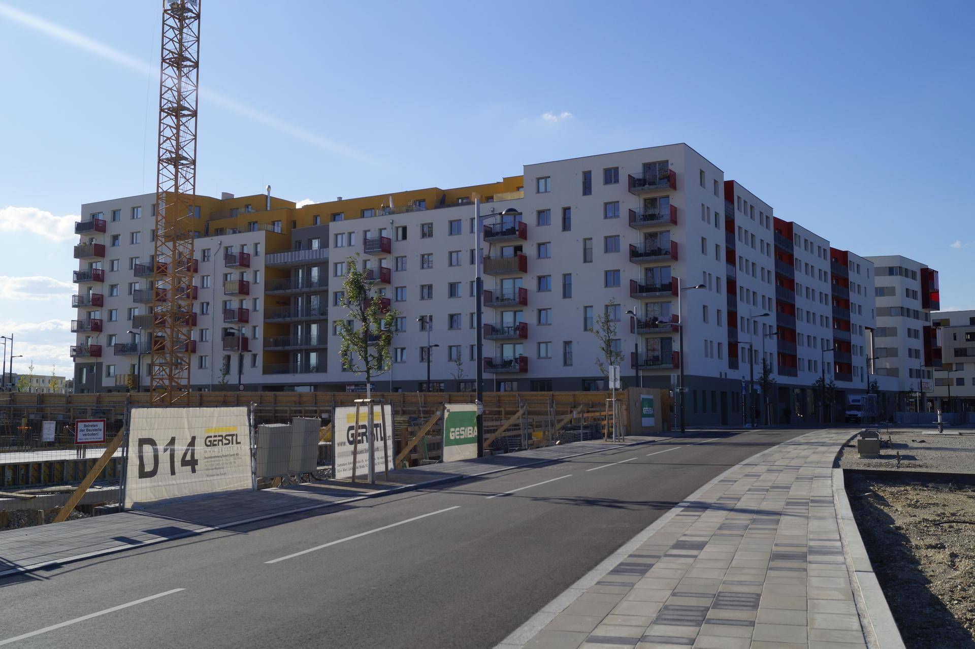 Großer Wohnkomplex im Bau mit moderner Architektur, roten Balkonen und einem Baukran im Hintergrund.