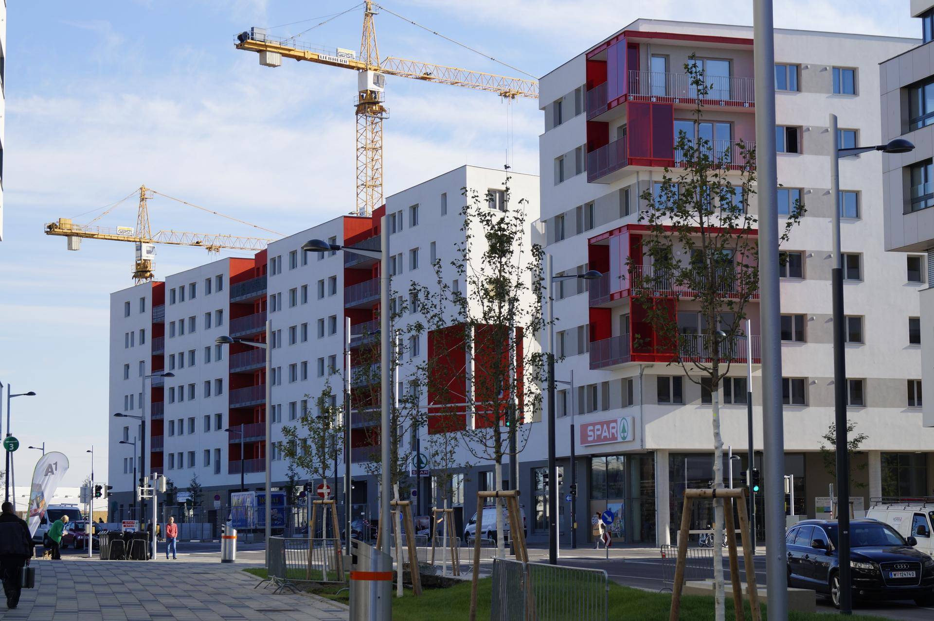 Moderne Wohn- und Geschäftsgebäude mit roten Balkonen und Ladenflächen im Erdgeschoss, belebtes Stadtbild.