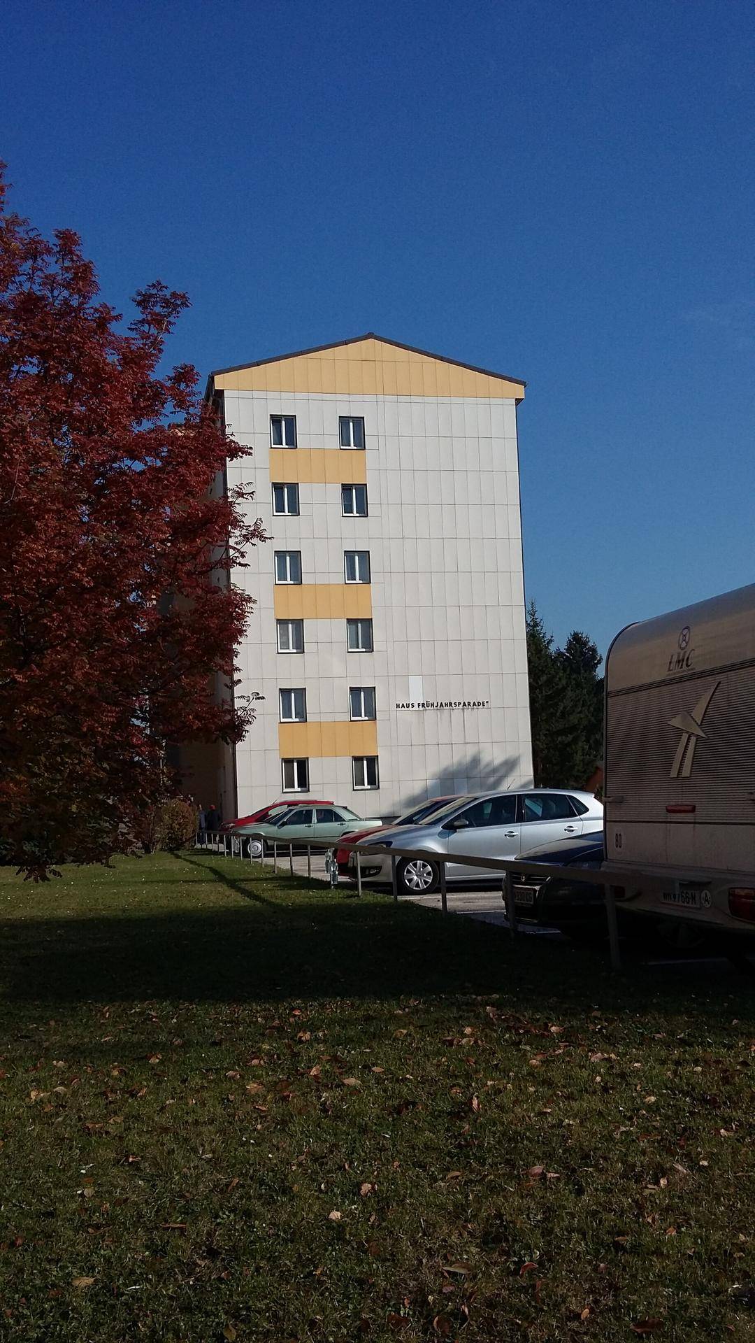 Seitenansicht eines Wohnblocks mit gelben Akzenten und parkenden Fahrzeugen auf einer Grünfläche.