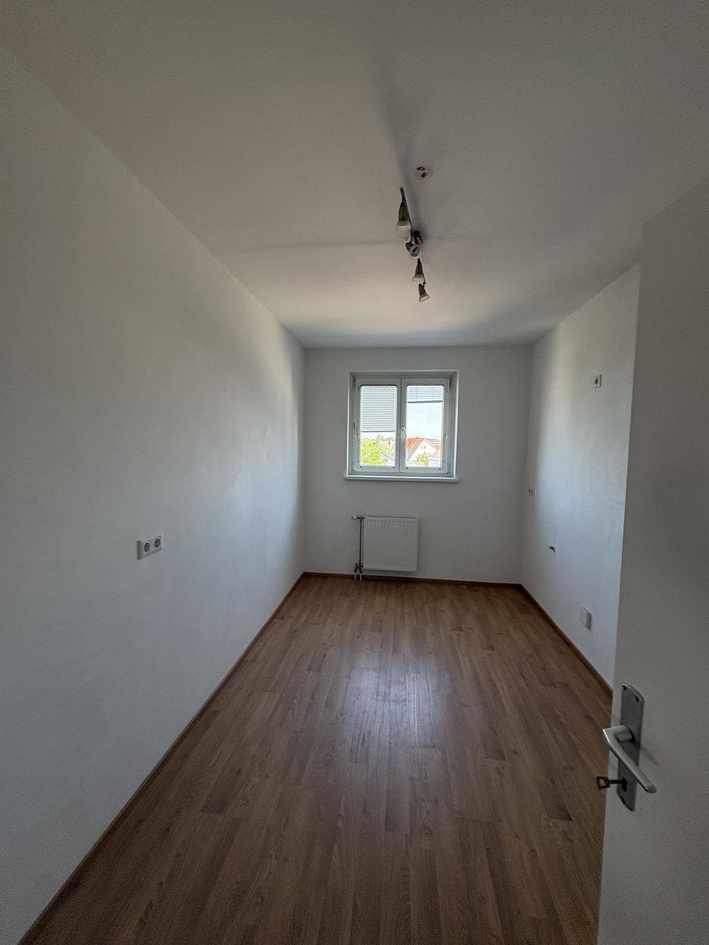 Leeres Zimmer mit Laminatboden, weißen Wänden und einem Fenster.