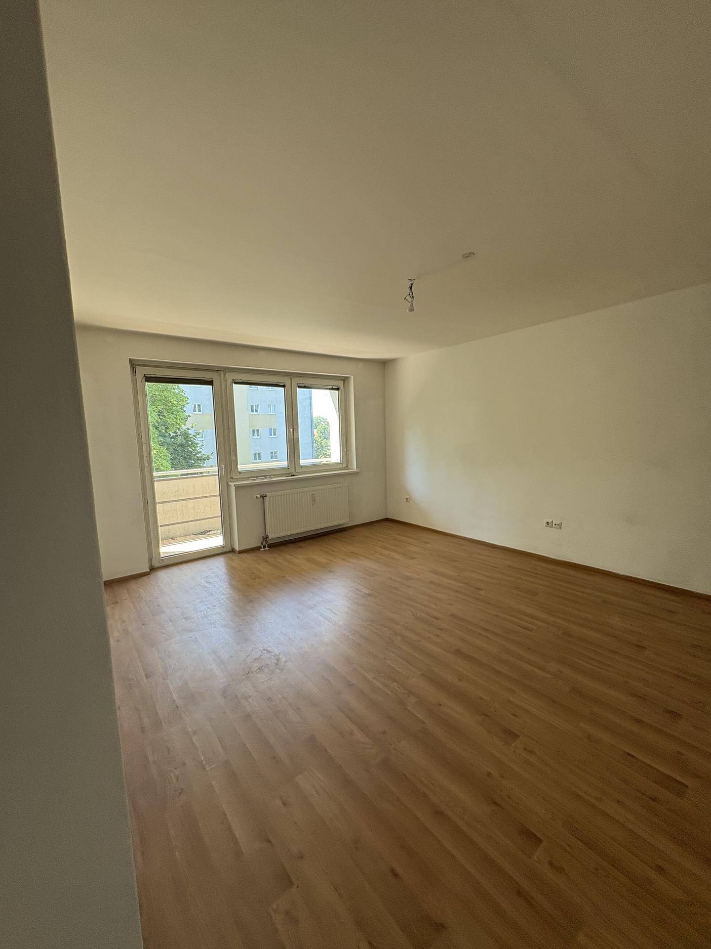 Helles Wohnzimmer mit Laminatboden, weißen Wänden und Zugang zum Balkon.