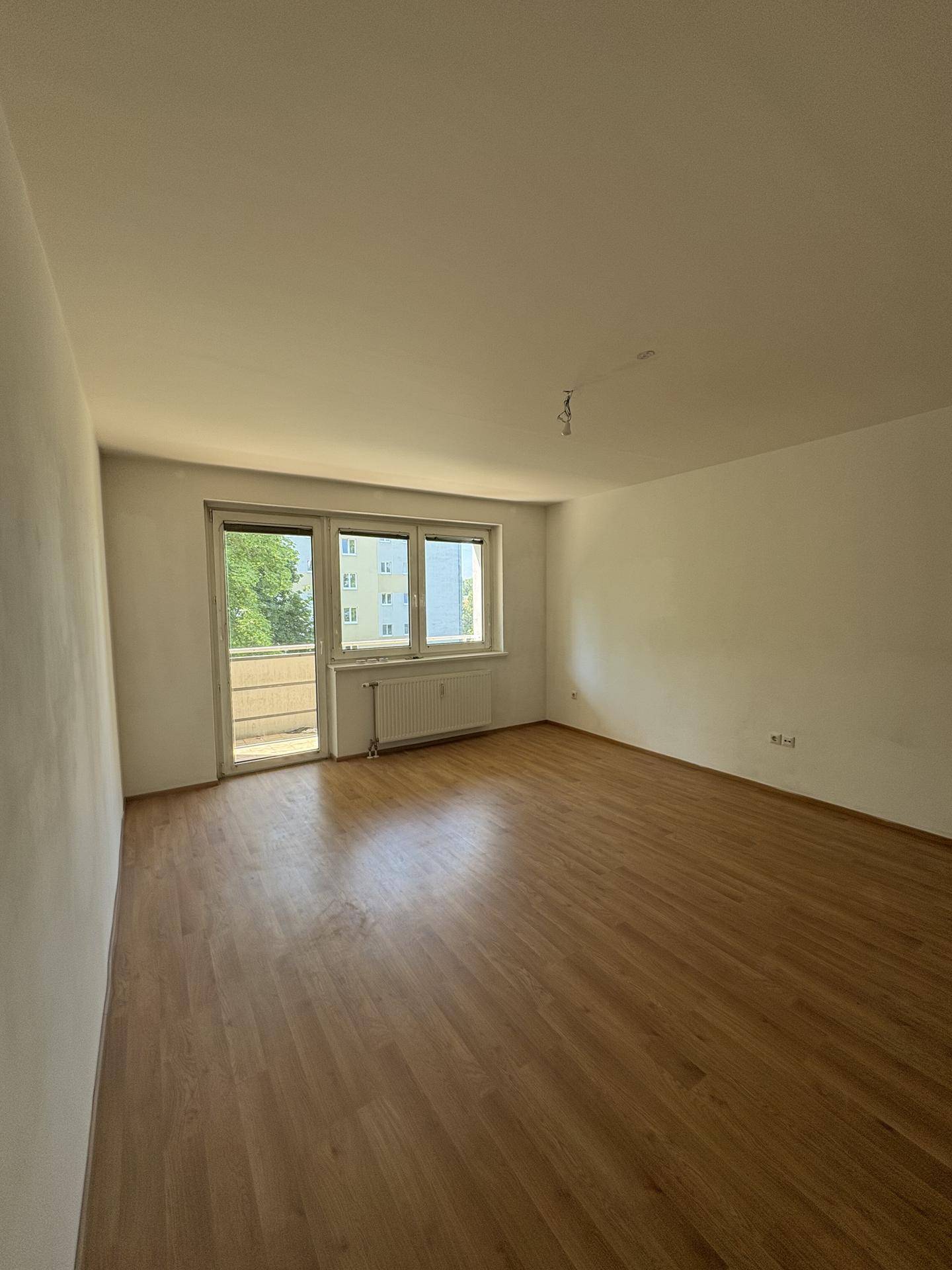 Lichtdurchflutetes Wohnzimmer mit Laminatboden und Zugang zum Balkon.