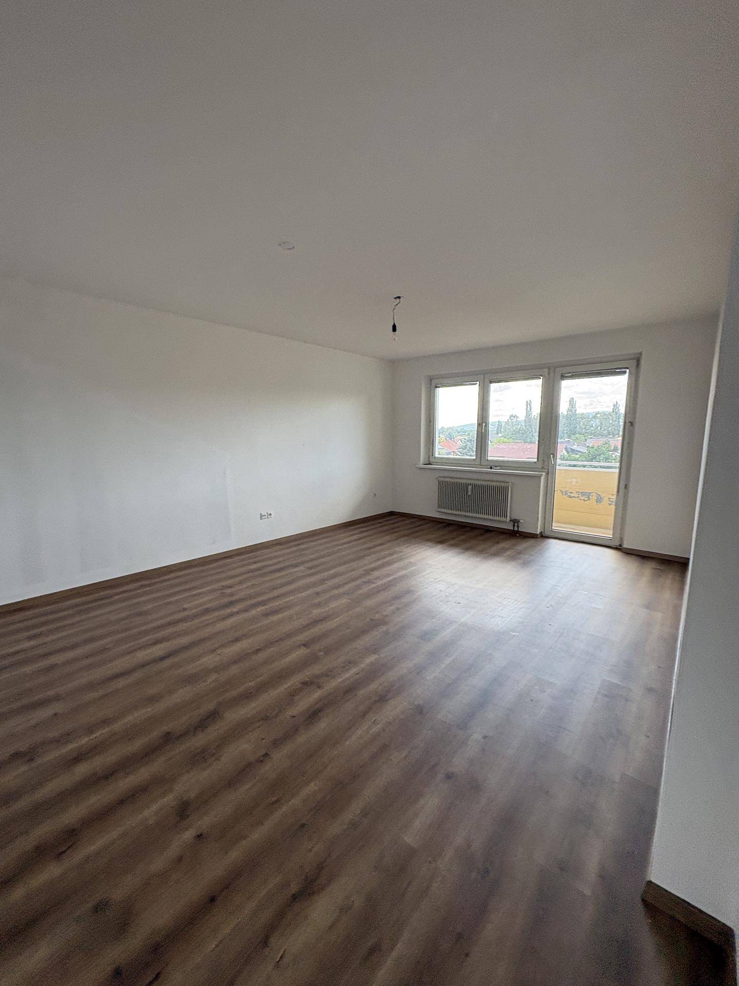 Geräumiges, leeres Wohnzimmer mit Laminatboden, weißen Wänden und Zugang zum Balkon.