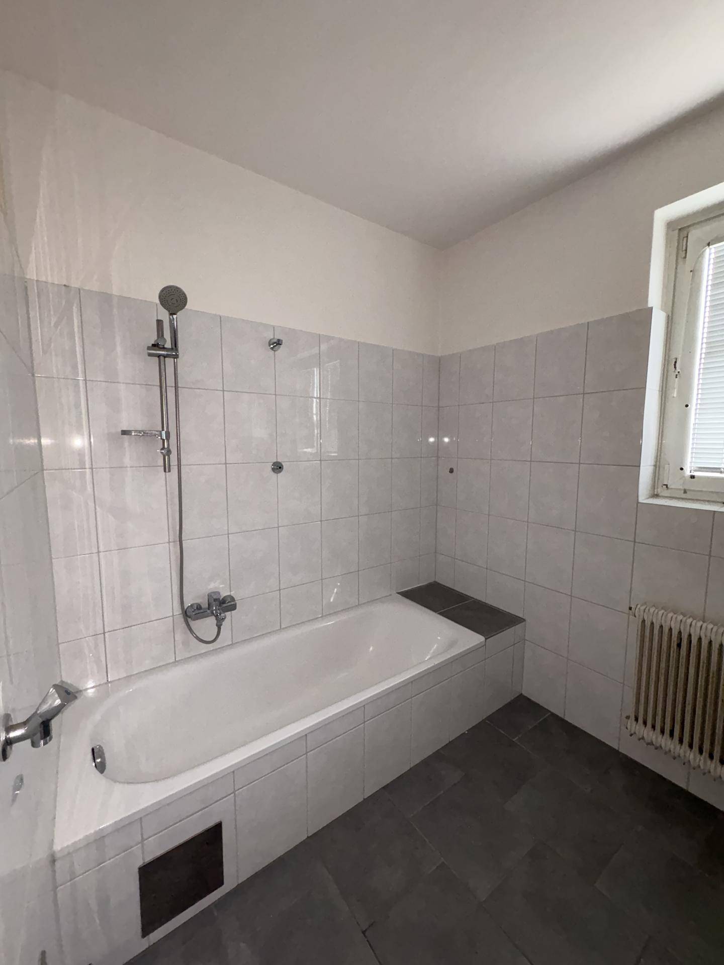 Badezimmer mit Badewanne, Duscharmatur, weißen Fliesen und dunklem Boden in gutem Zustand.