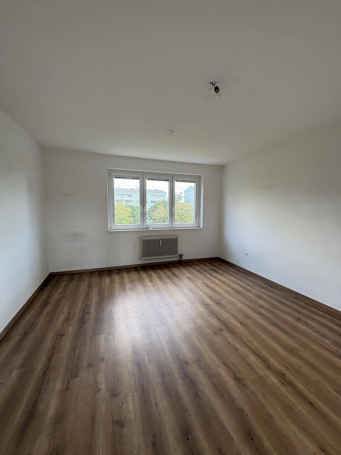 Leeres Zimmer mit Laminatboden, weißen Wänden und großem Fenster mit Blick ins Grüne.