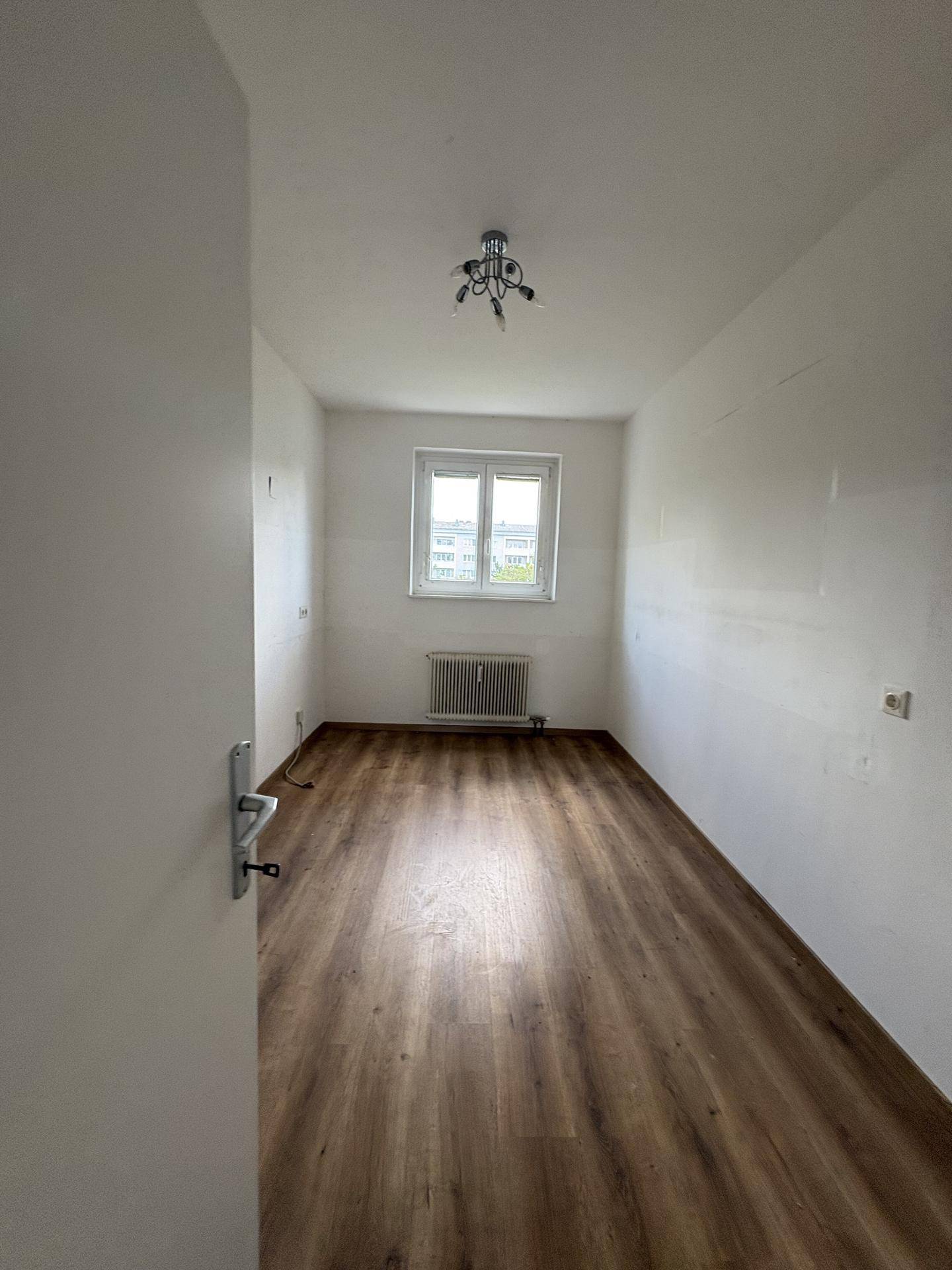 Langes, schmales Zimmer mit Laminatboden, weißen Wänden und einem Fenster für Tageslicht.